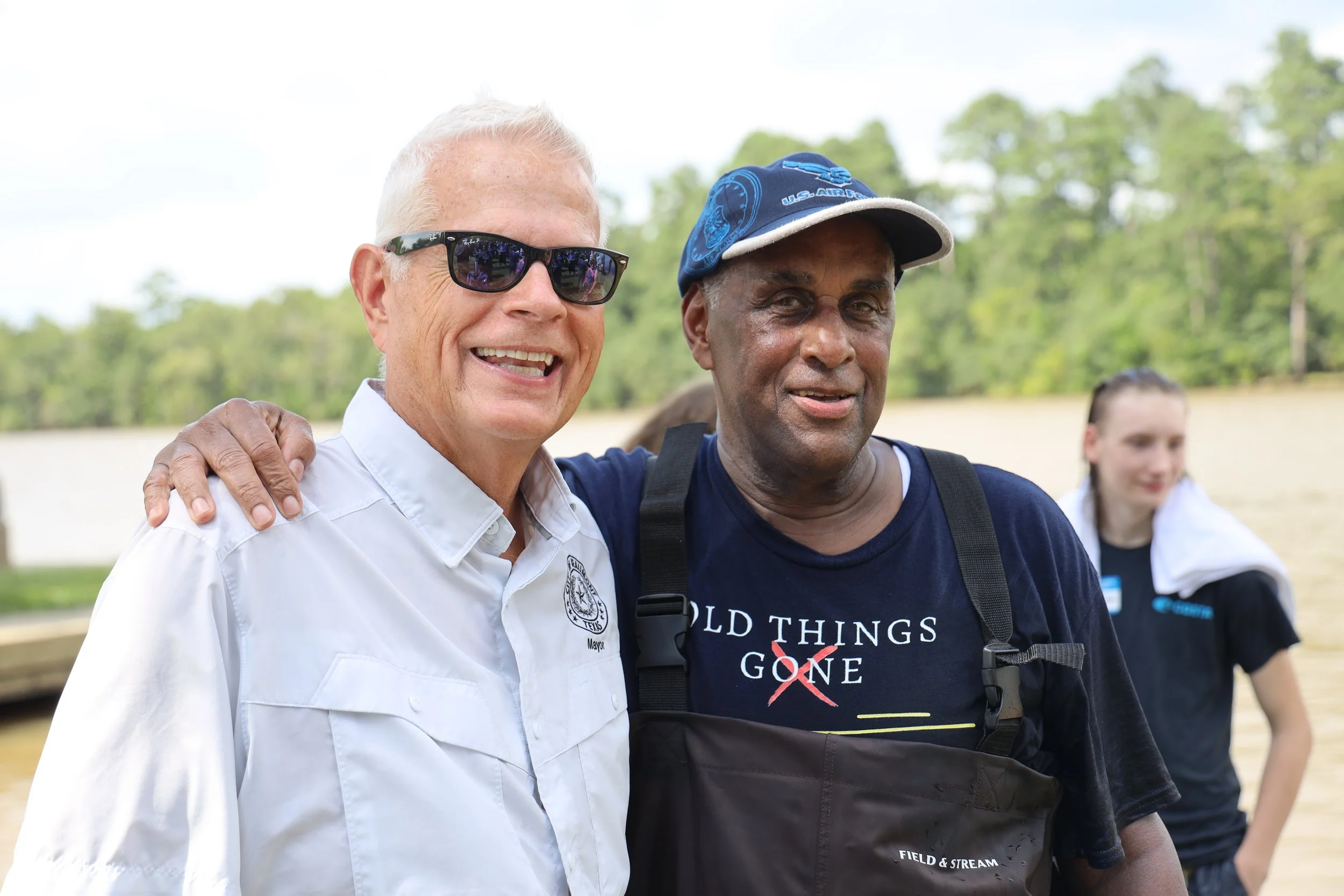 Two men smiling and posing outdoors on a sunny day. One man has white hair, is wearing glasses, a white shirt with an emblem, and has his hand on the other man's shoulder. The other man has dark skin, is wearing a dark t-shirt with writing, and a blue cap. In the background, a woman with light hair is slightly out of focus, standing amidst greenery.