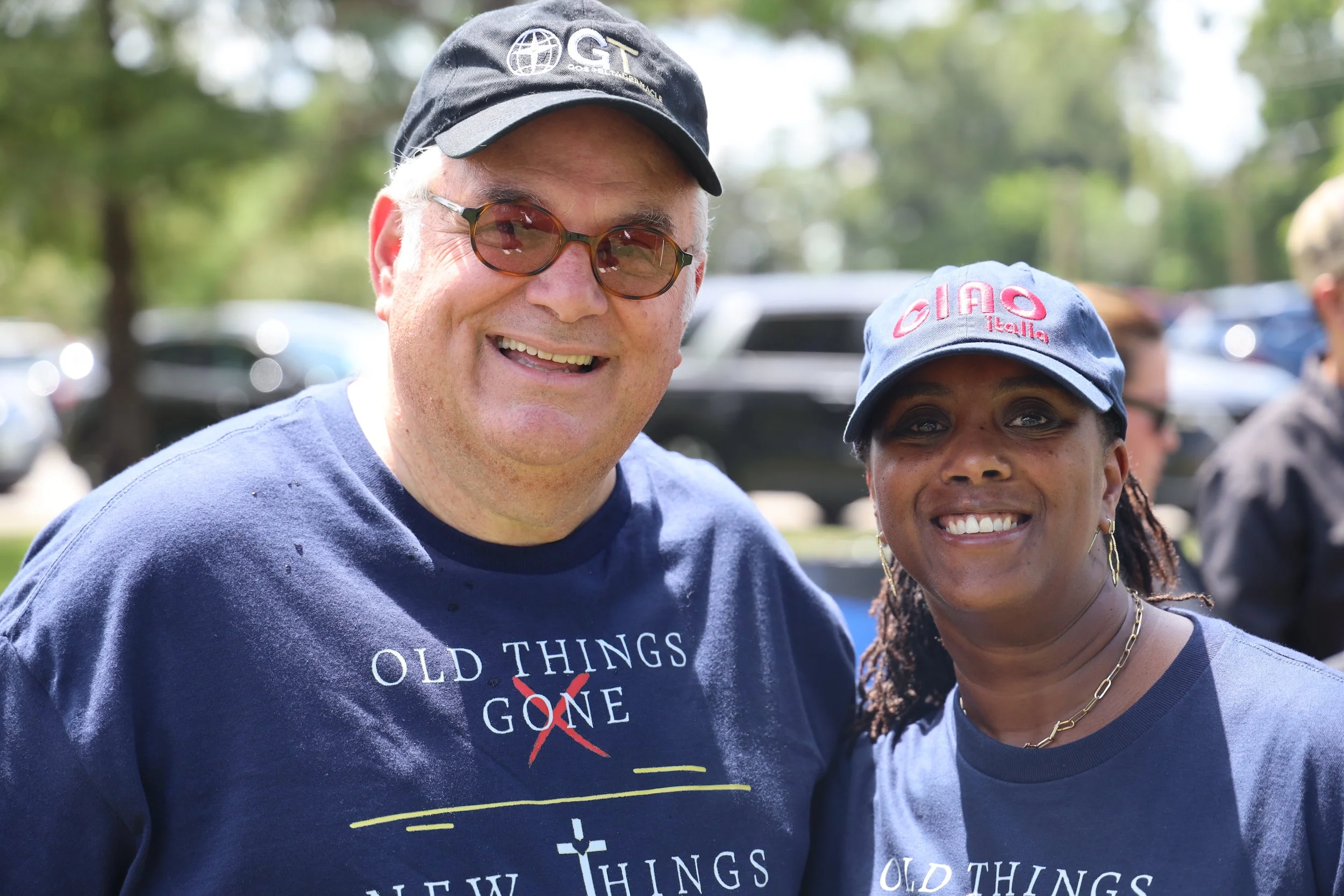 Two smiling people are outdoors, a man wearing sunglasses, a black cap with a logo, and a blue t-shirt, and a woman wearing a blue cap with red text, earrings, and a gray t-shirt.