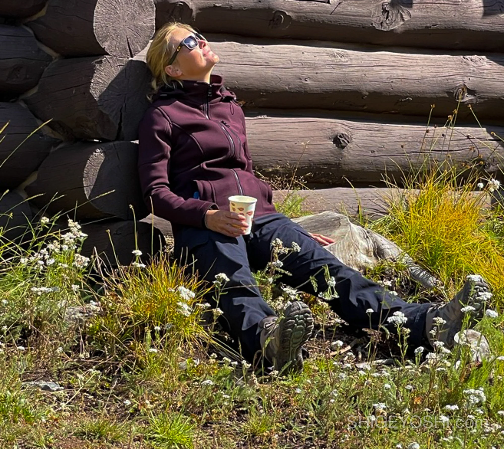A woman with blonde hair, wearing a purple jacket and sunglasses, is sitting outdoors against a wooden log wall. She is holding a cup and relaxing in sunlight with her eyes closed.