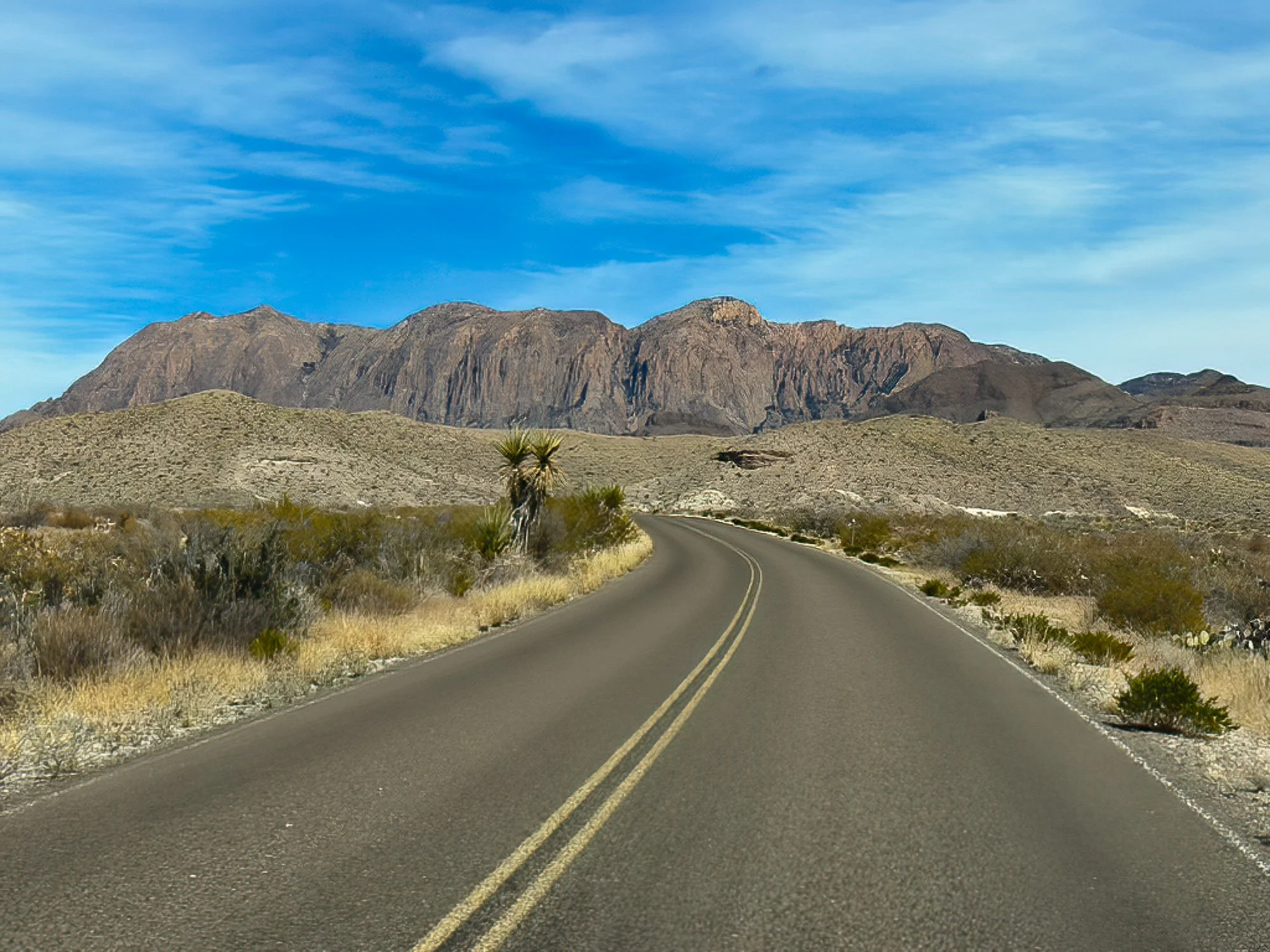 Two-lane desert road curving toward rugged mountain peaks under a blue West Texas sky