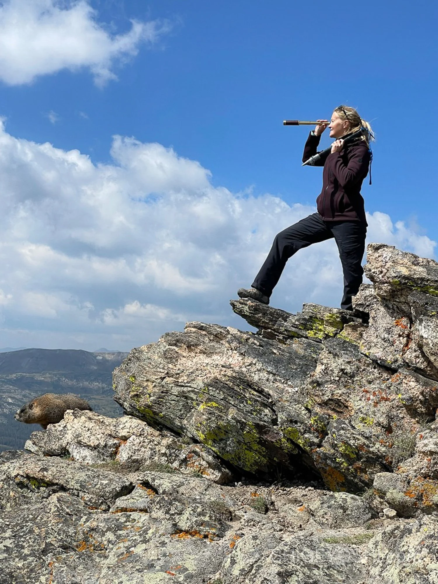 A woman standing on a rocky mountain ledge, looking through a telescope at the mountains and sky in the distance.