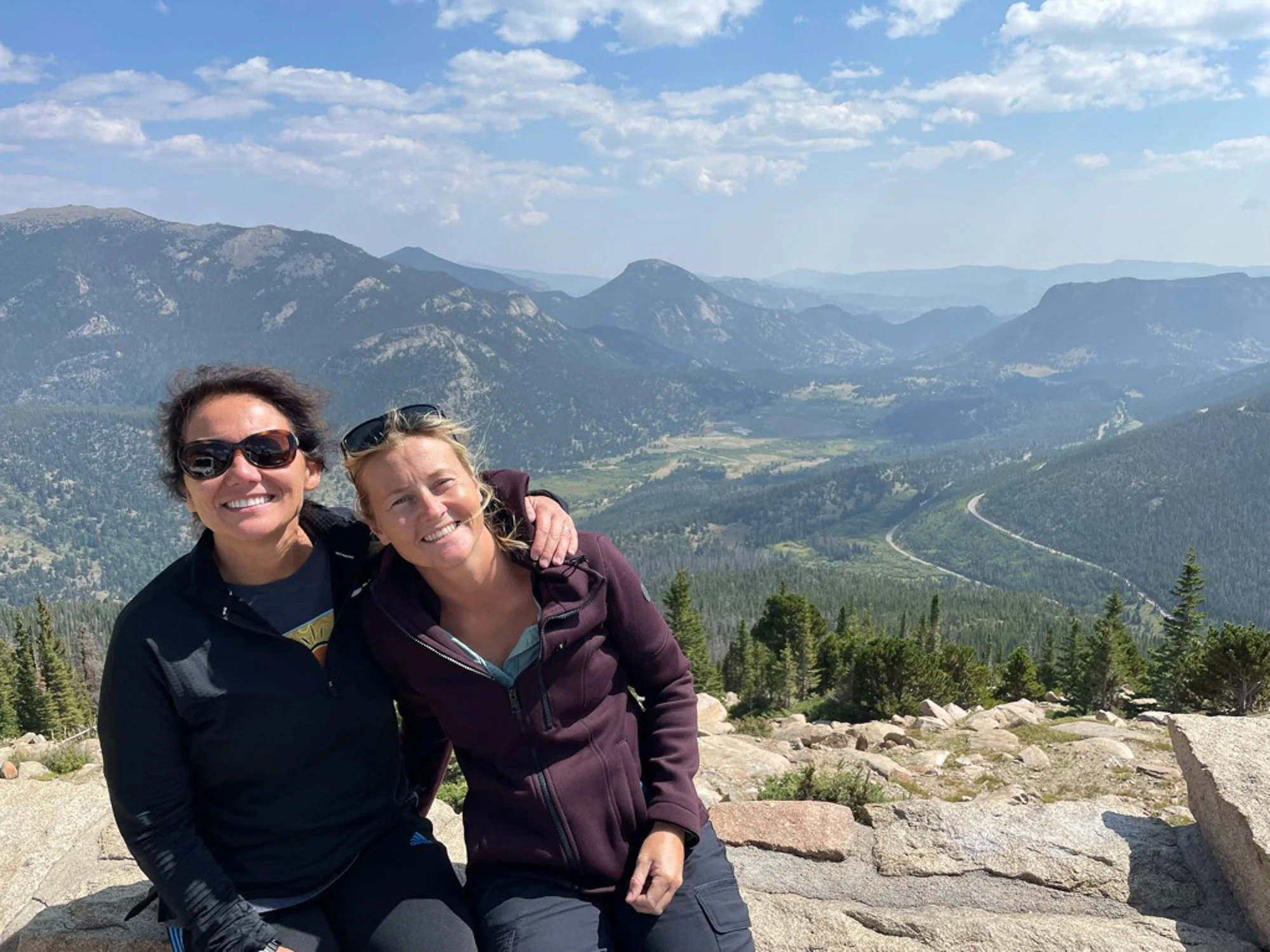 wo women smiling together at a mountain overlook with a sweeping forested valley and winding road below