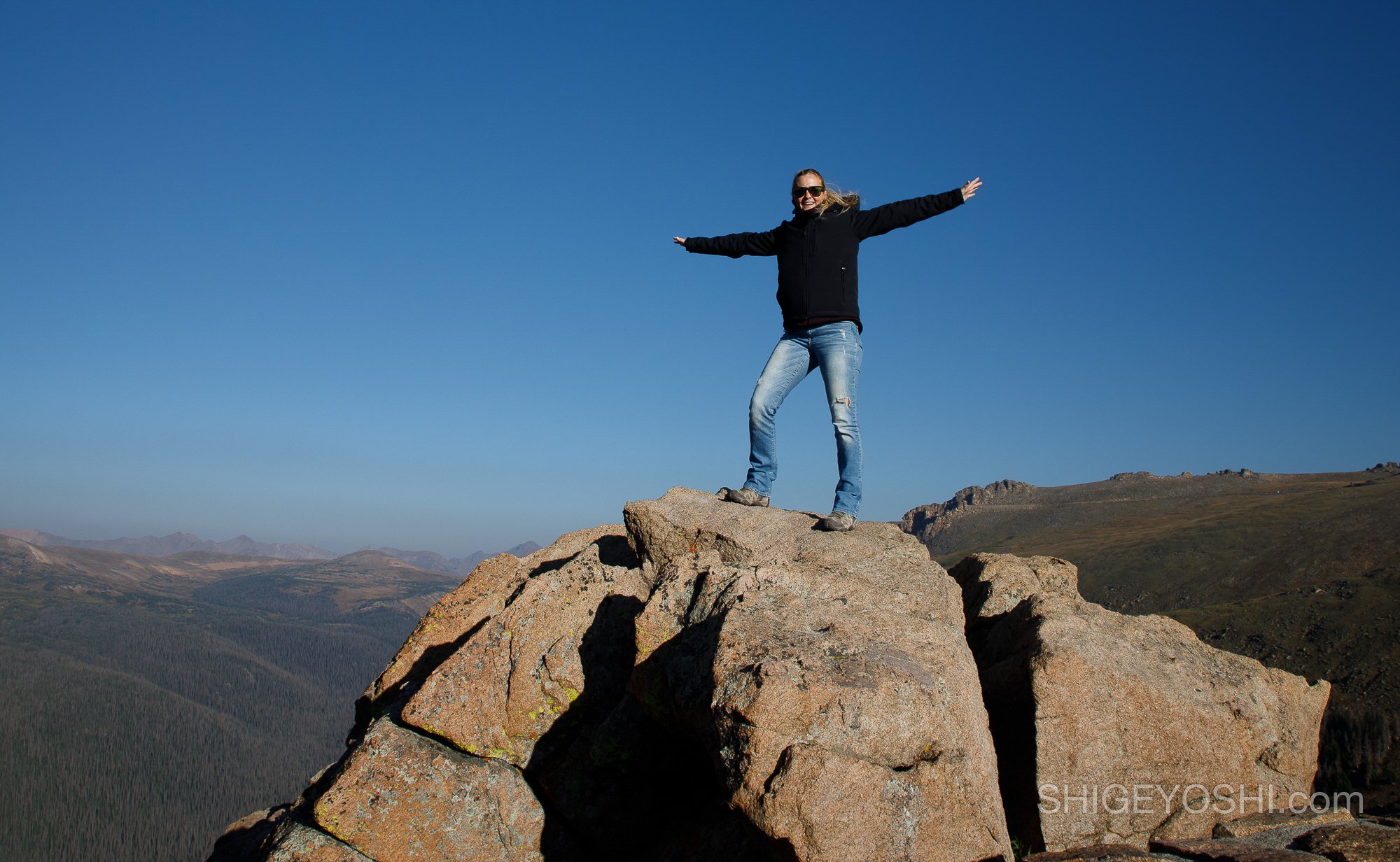 A woman standing on top of a large rock formation with her arms outstretched, wearing sunglasses, a black jacket, and jeans, with a vast mountain landscape in the background under a clear blue sky.