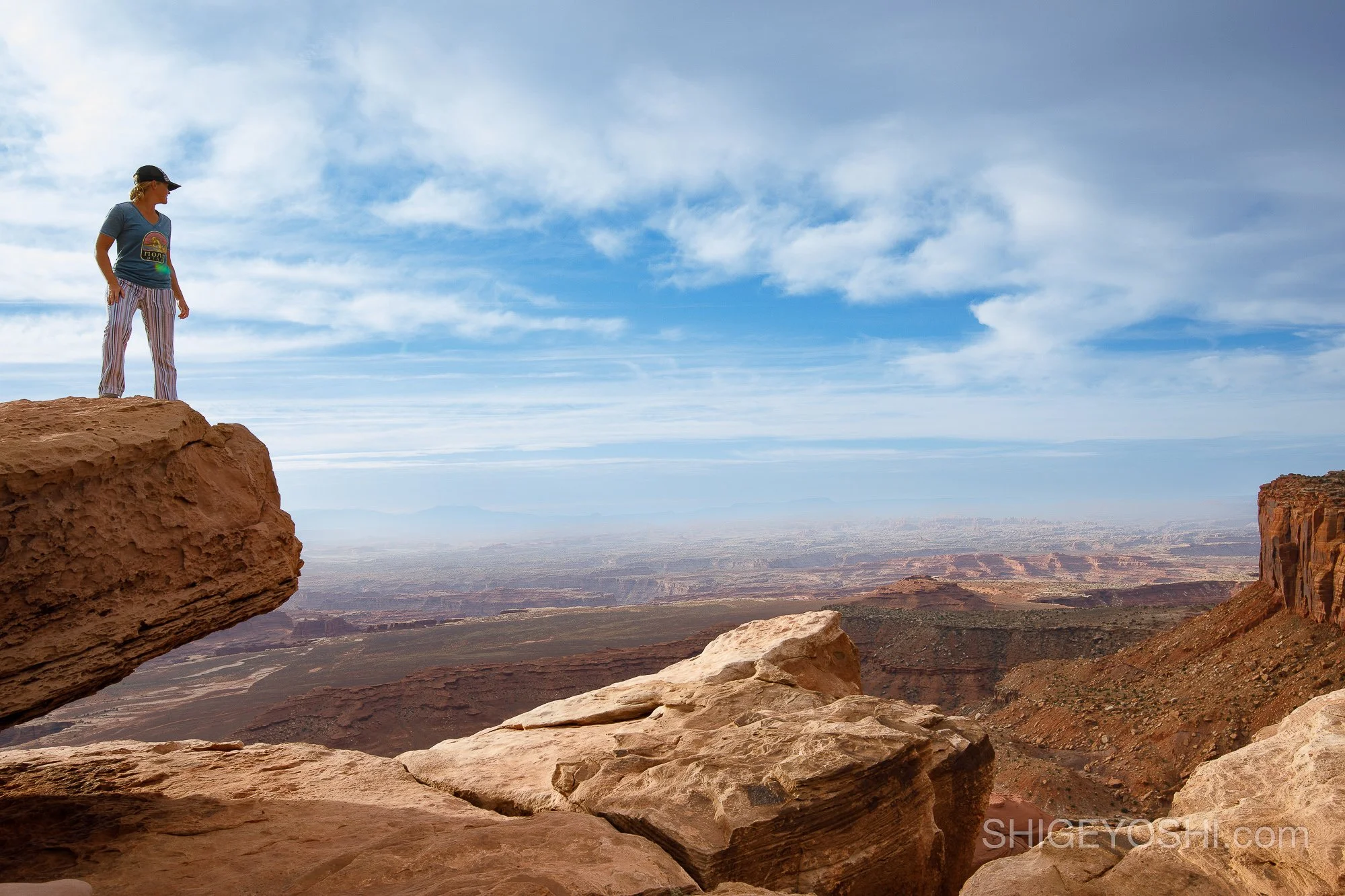 A woman standing on a large rock ledge overlooking a vast desert landscape with rock formations under a partly cloudy sky.