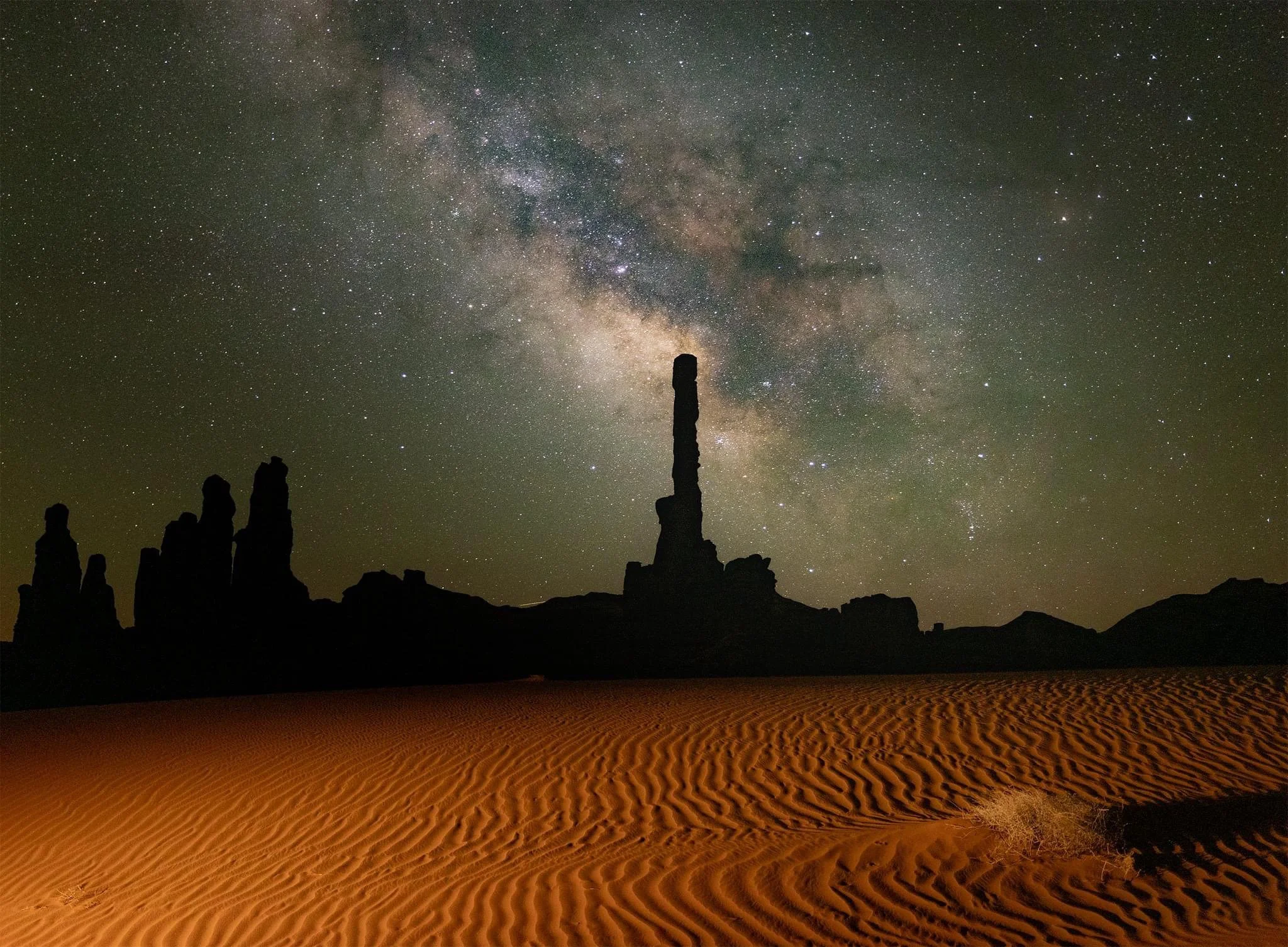 Night sky filled with stars and the Milky Way, with rock formations in the foreground and sand dunes at the bottom.
