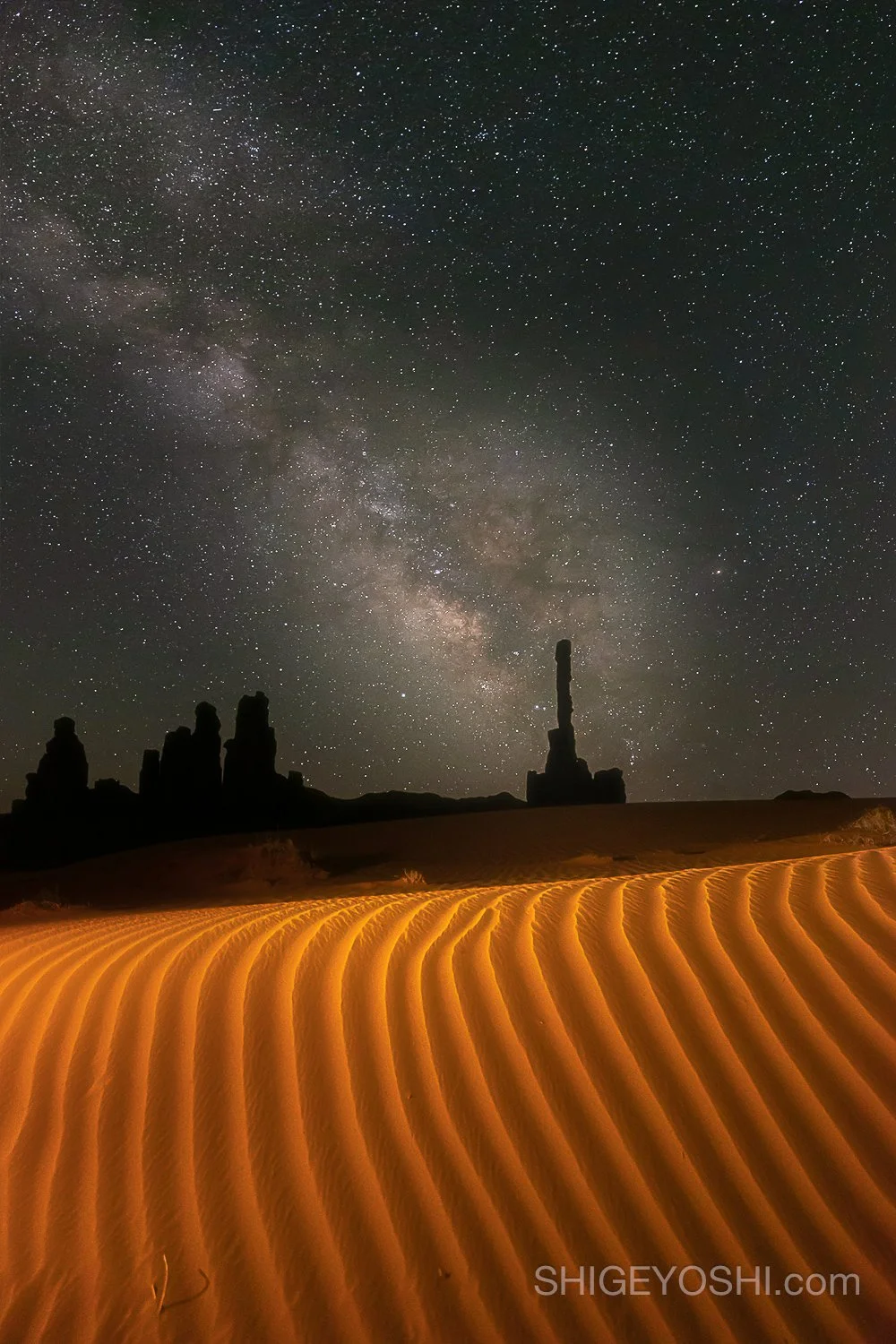 Night sky filled with stars and the Milky Way galaxy, desert landscape with sand dunes in the foreground, and rock formations in the distance.
