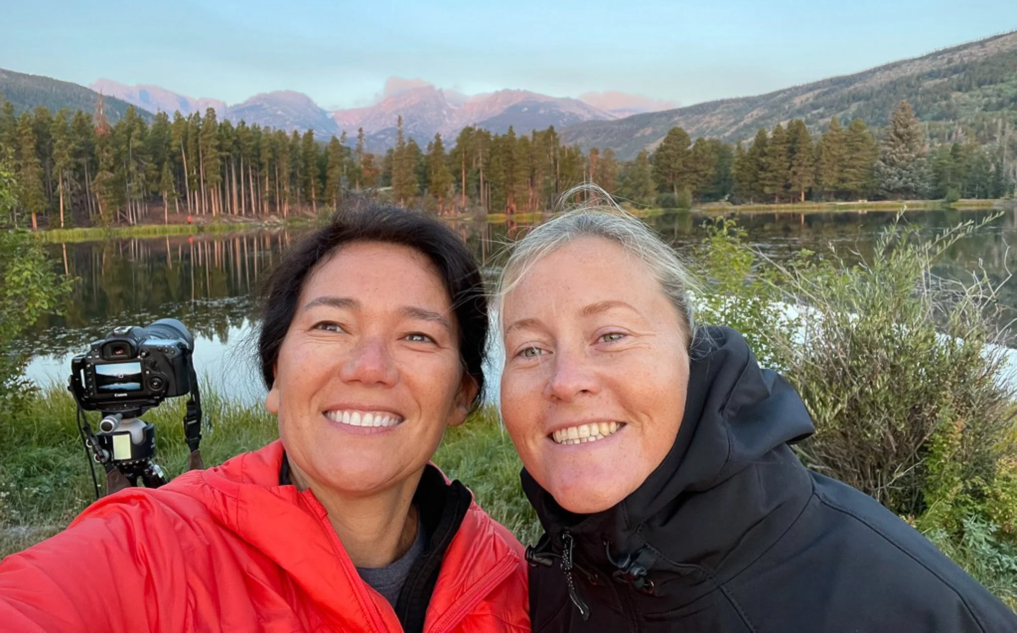 Two women smiling and taking a selfie by a lake with trees and mountains in the background during daylight.
