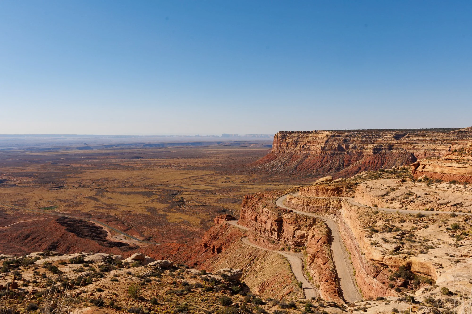 A narrow switchback road carved into red canyon walls in the American Southwest, with a vast desert landscape stretching to the horizon.