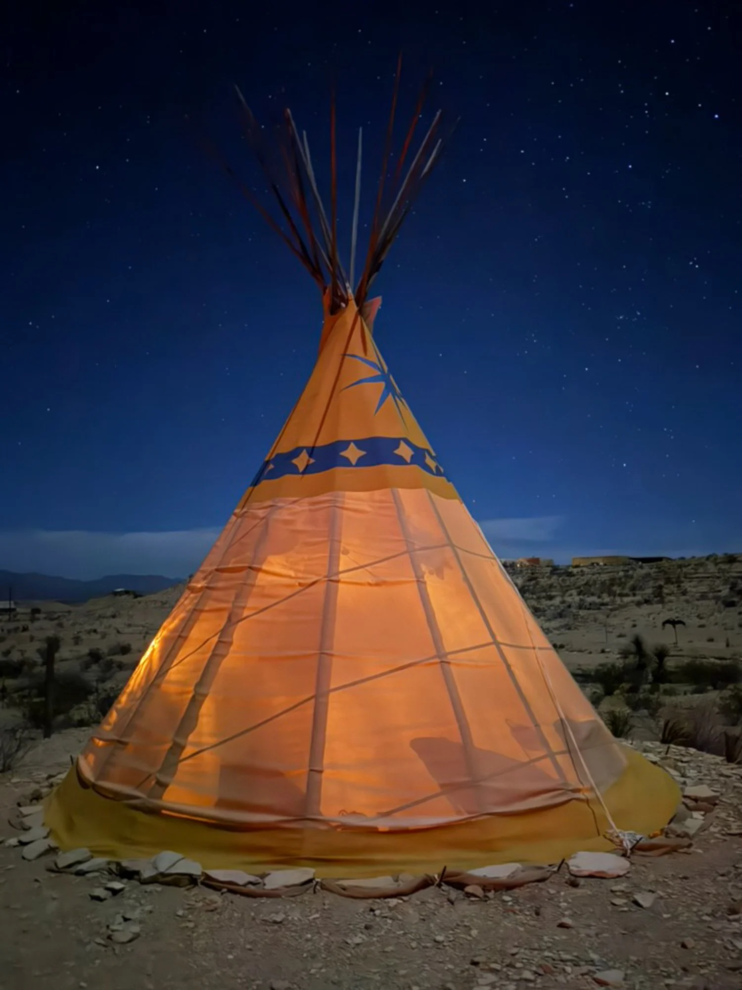 Illuminated teepee glowing orange against a deep blue night sky filled with stars in the Terlingua desert