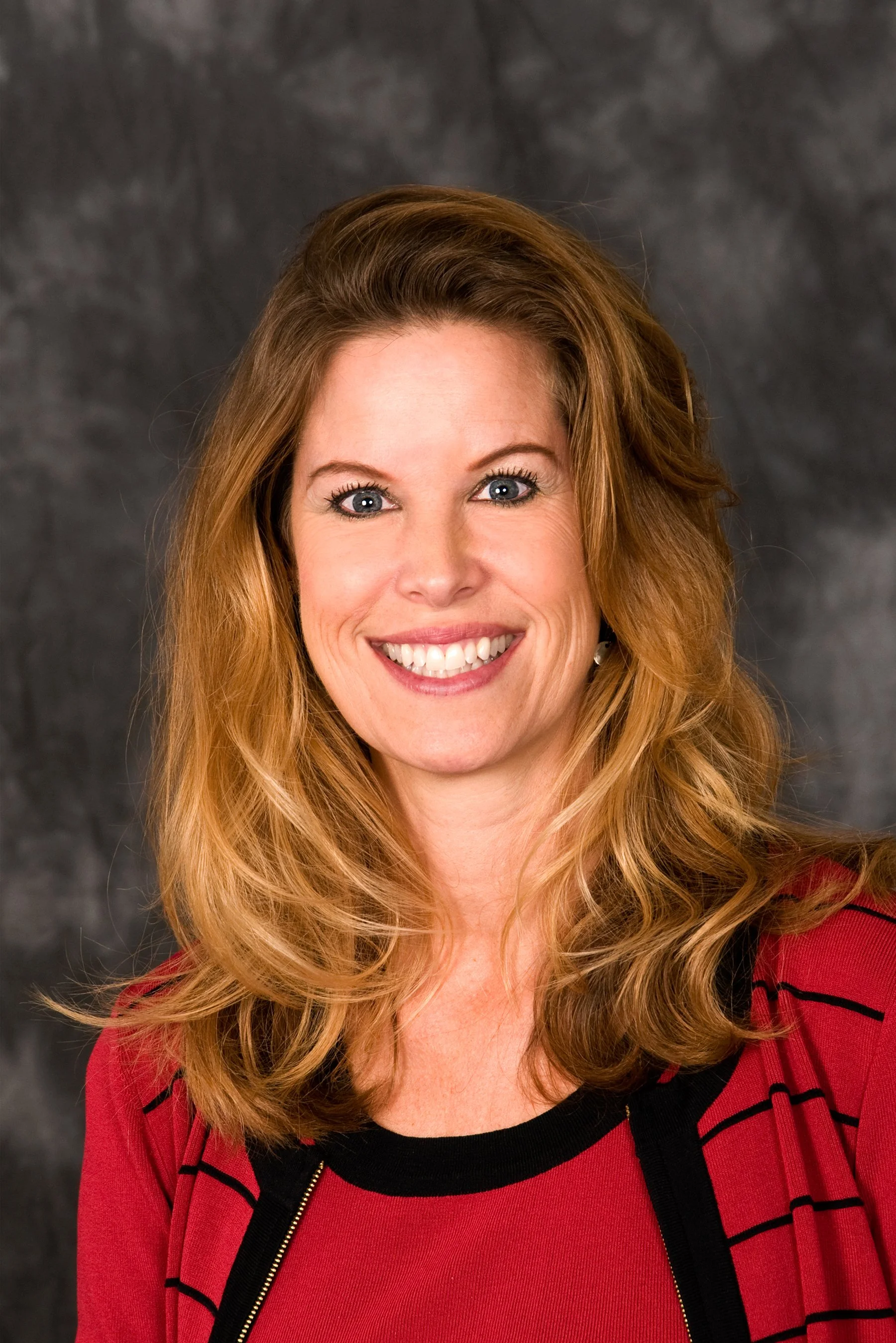 Portrait of a Laure Reed, CEO of RJL & Associates with wavy, light brown hair, smiling in front of a dark, textured background. She is wearing a red top with black accents.