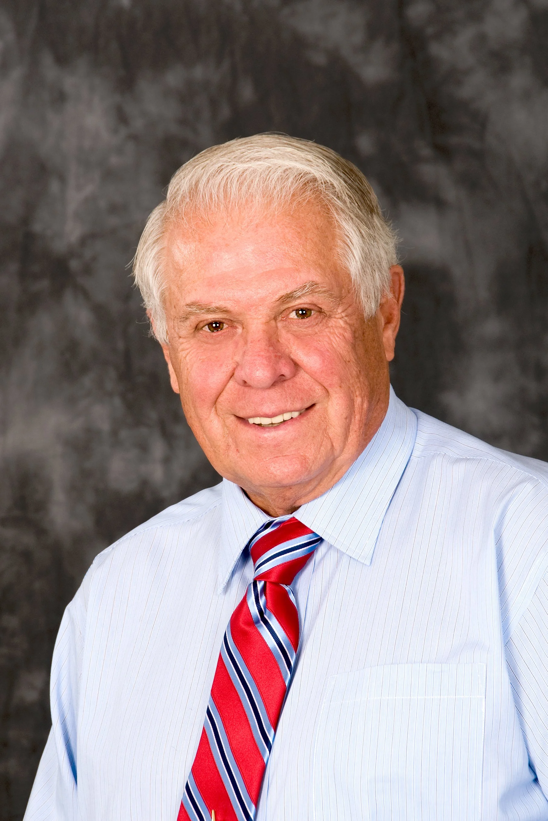 Robert J. Lung, founder of RJL & Associates, smiling with gray hair wearing a light blue shirt and a red, white, and blue striped tie against a dark mottled background.