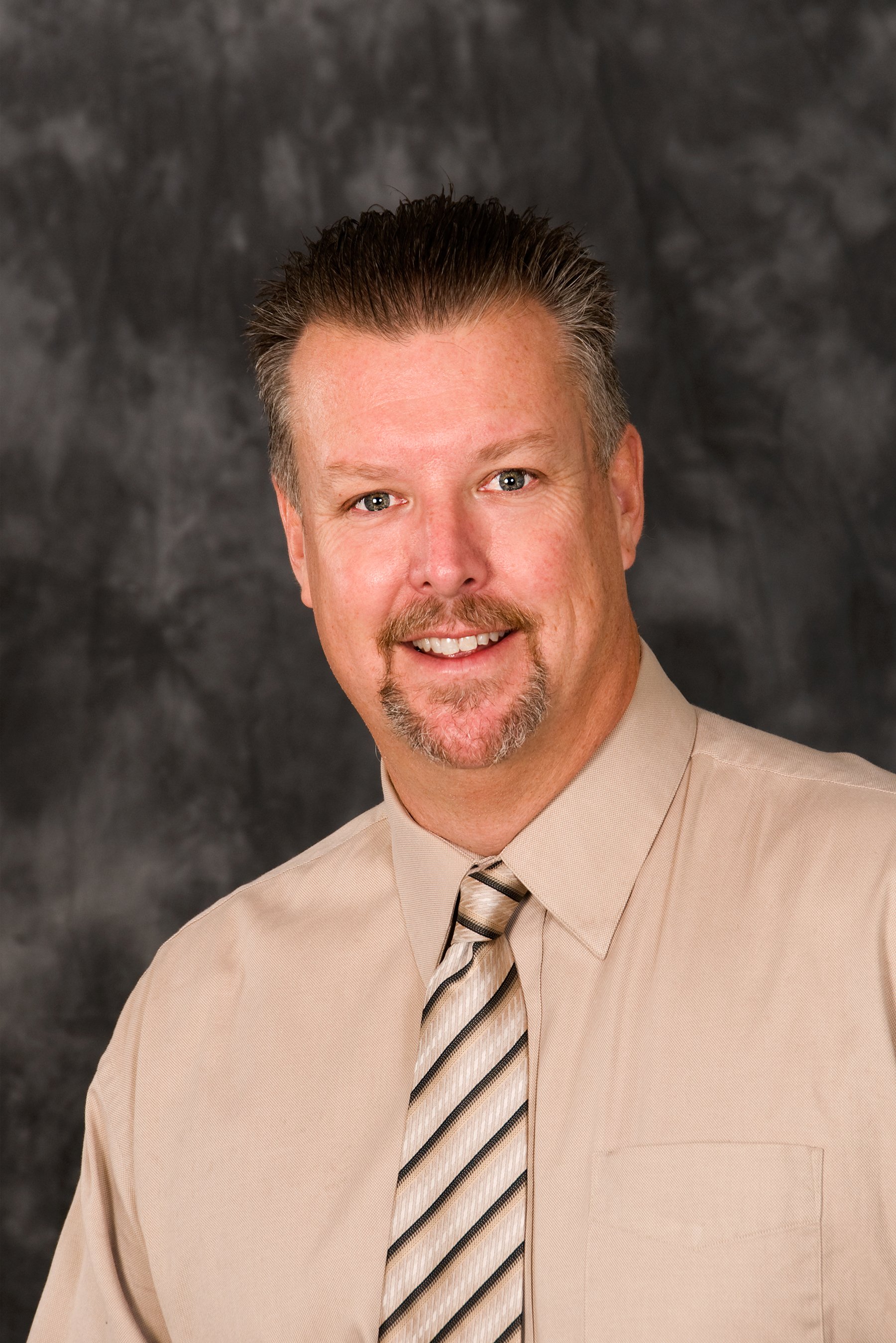 Headshot of Chris Lung, President of RJL & Associates, with short, spiked brown hair, blue eyes, and a goatee, wearing a beige dress shirt and a diagonally striped beige and brown tie, against a dark, textured background.