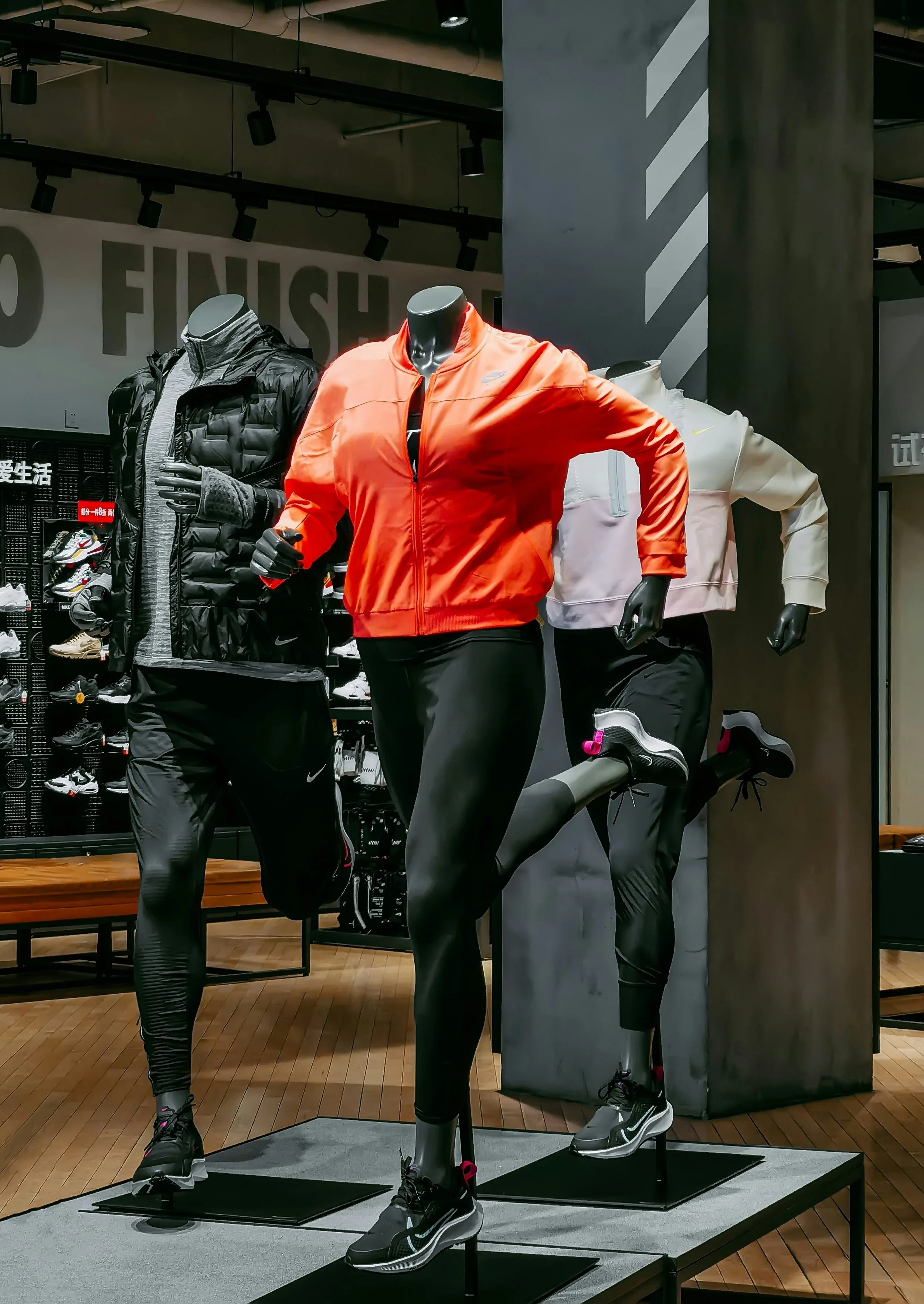 Display of athletic apparel including a black puffer jacket, gray shirt, orange Nike jacket, white Nike jacket, black leggings, and running shoes on mannequins in a store.