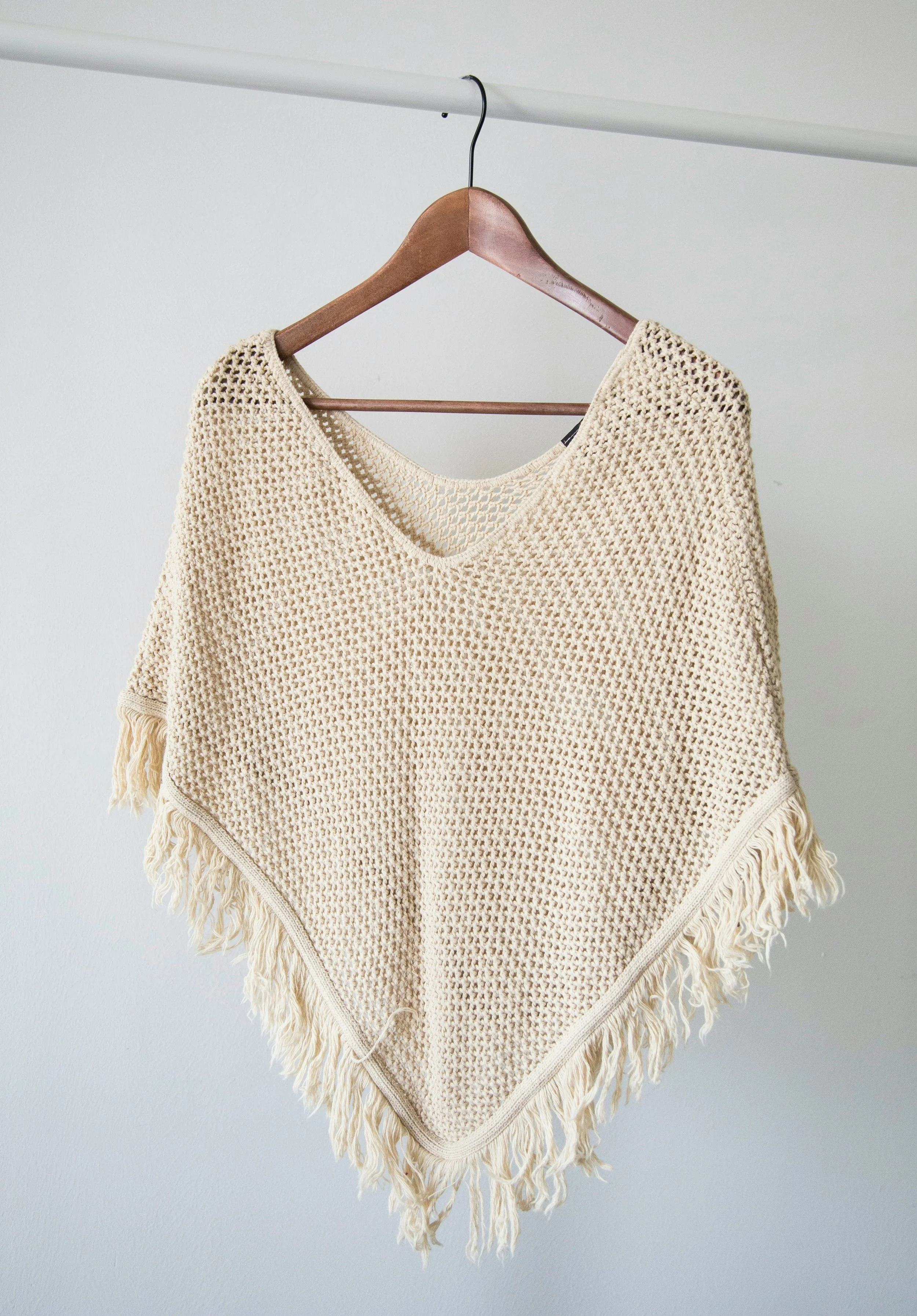 Beige crocheted poncho with fringe hanging on a wooden hanger, suspended from a metal rod against a plain white wall.