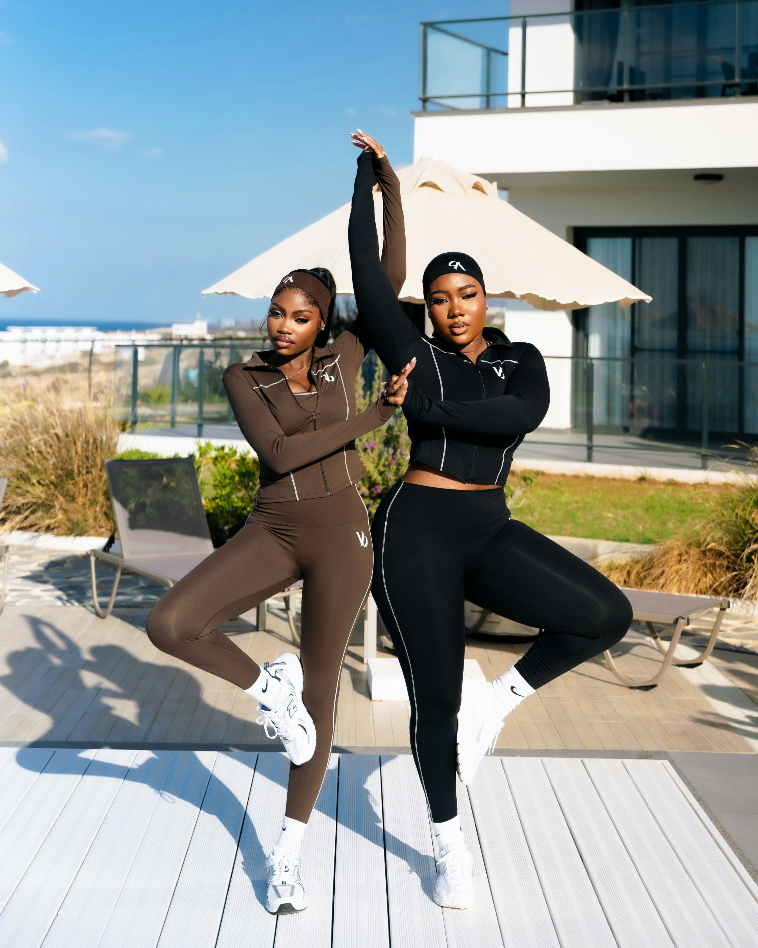 Two women in athletic outfits and headbands practicing fitness poses outdoors on a balcony with white umbrellas and modern building in the background.