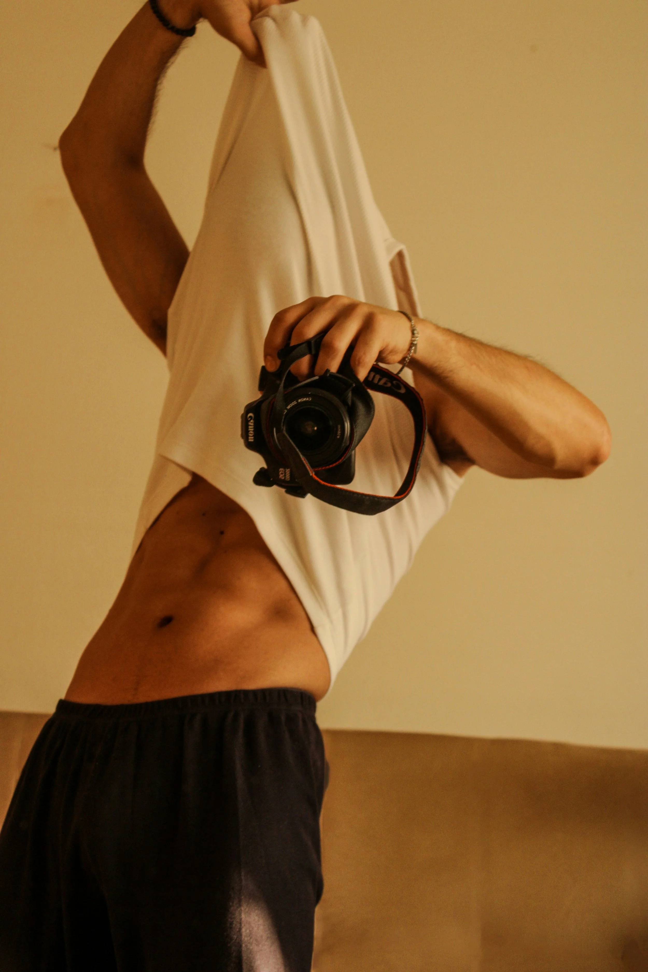 Person taking a mirror selfie with a Canon camera, wearing a white T-shirt and black pants, holding shirt in front of face, standing against beige wall.