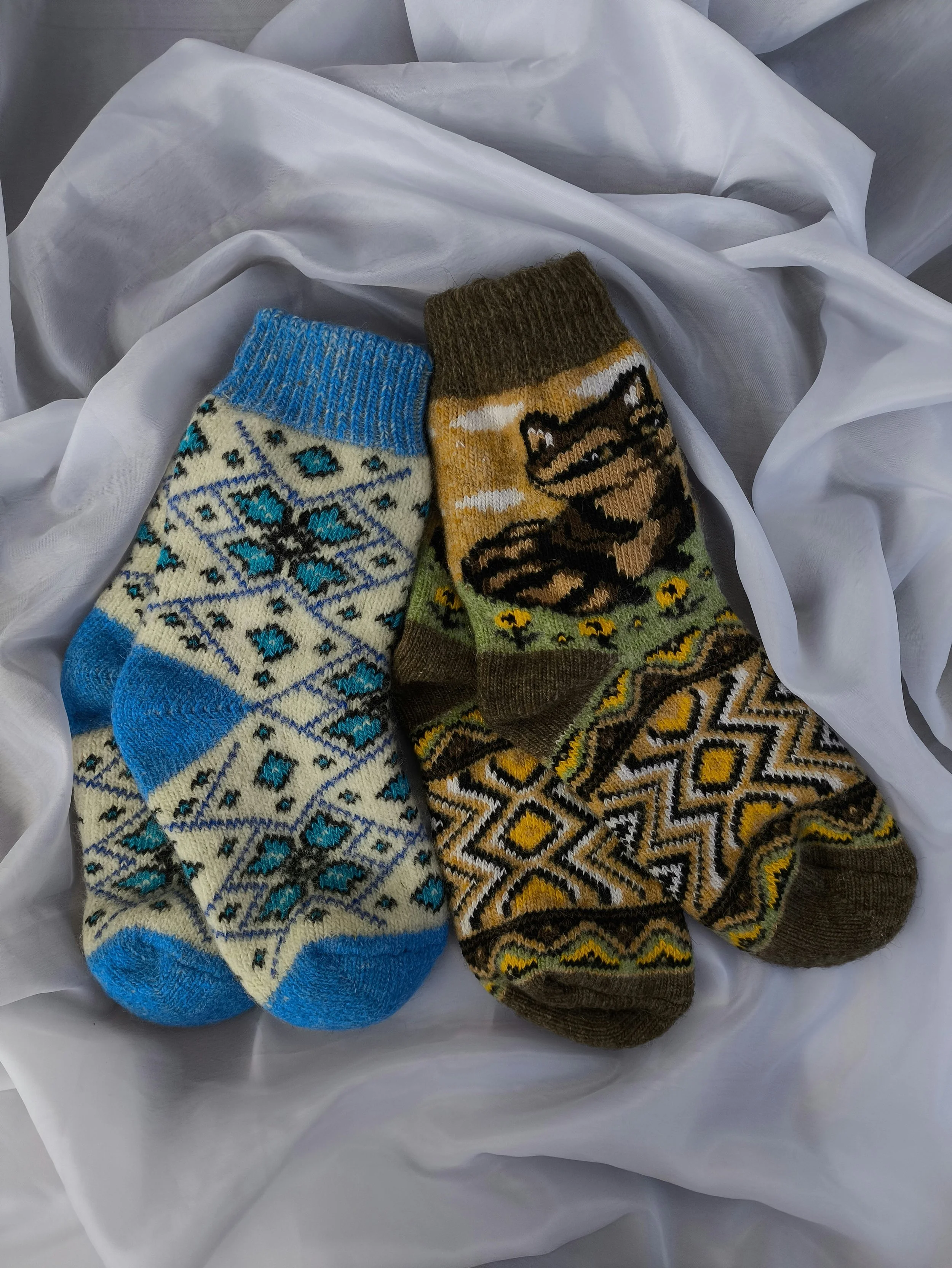 Two colorful knitted socks with geometric patterns, one in blue and cream with diamond shapes, and the other in brown, yellow, black, and green with a zigzag pattern, resting on crumpled white fabric.