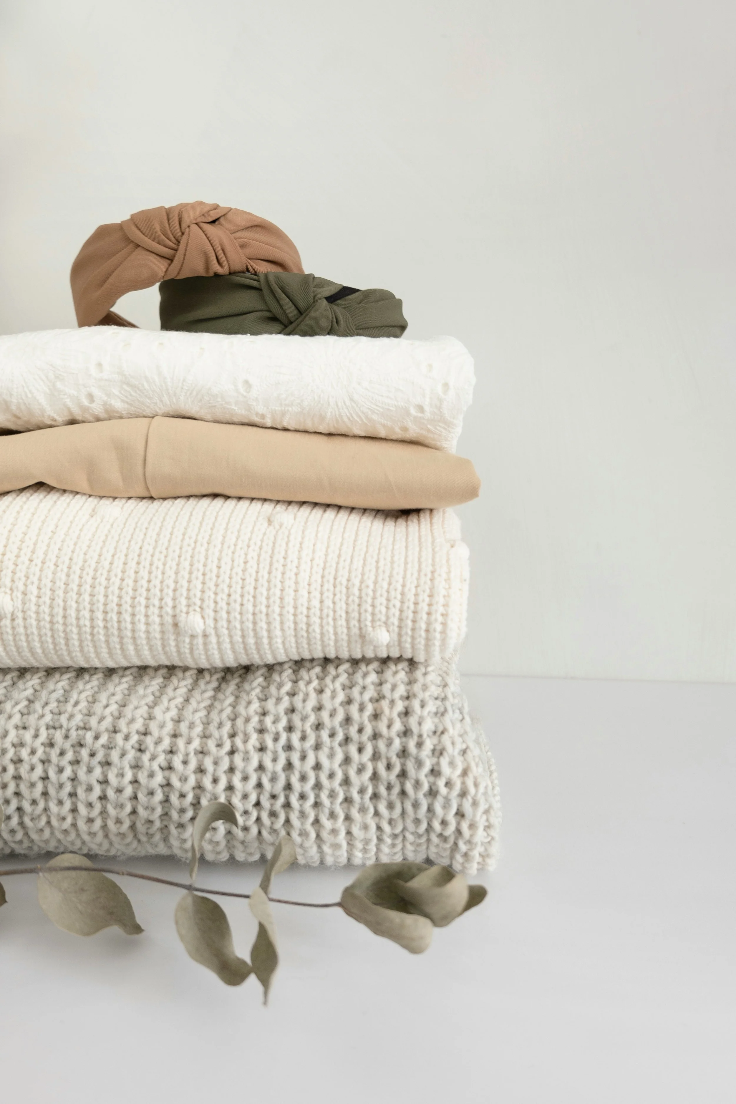 Stack of folded knit blankets in neutral tones with dried eucalyptus leaves in front, against a plain white background.