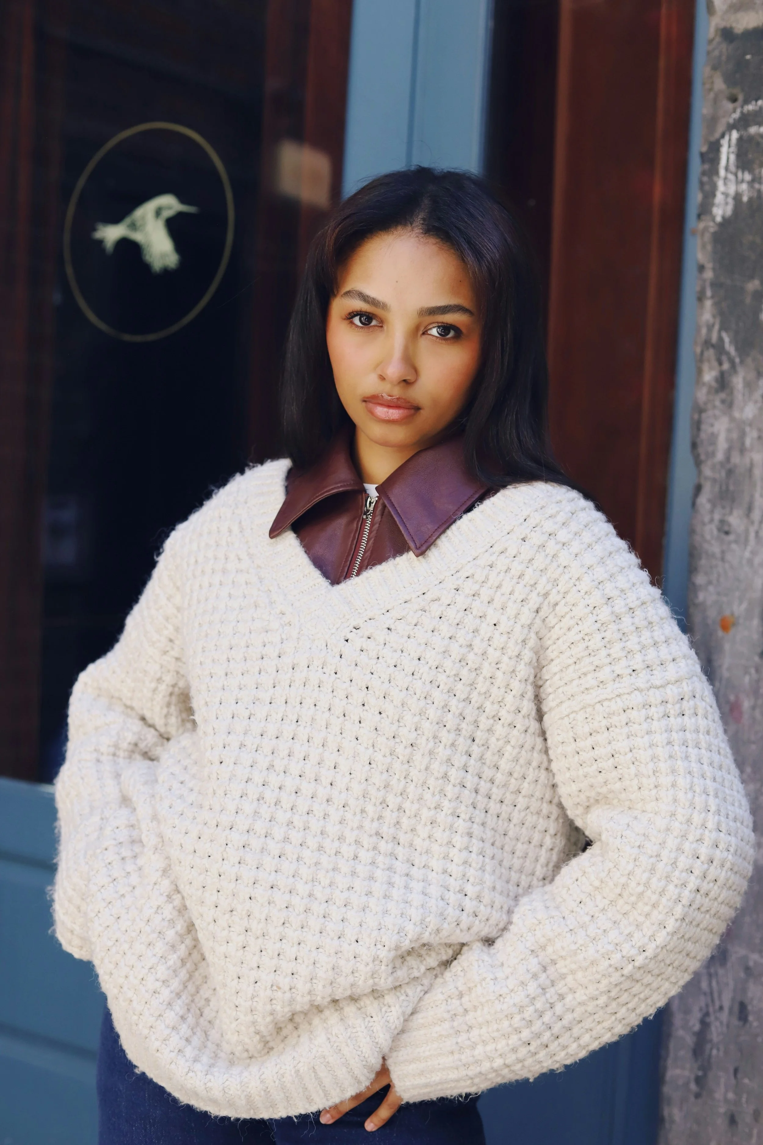 A woman with black hair and brown skin wearing a white knit sweater with a brown leather collar, standing outdoors near a stone wall and a door with a hummingbird design.
