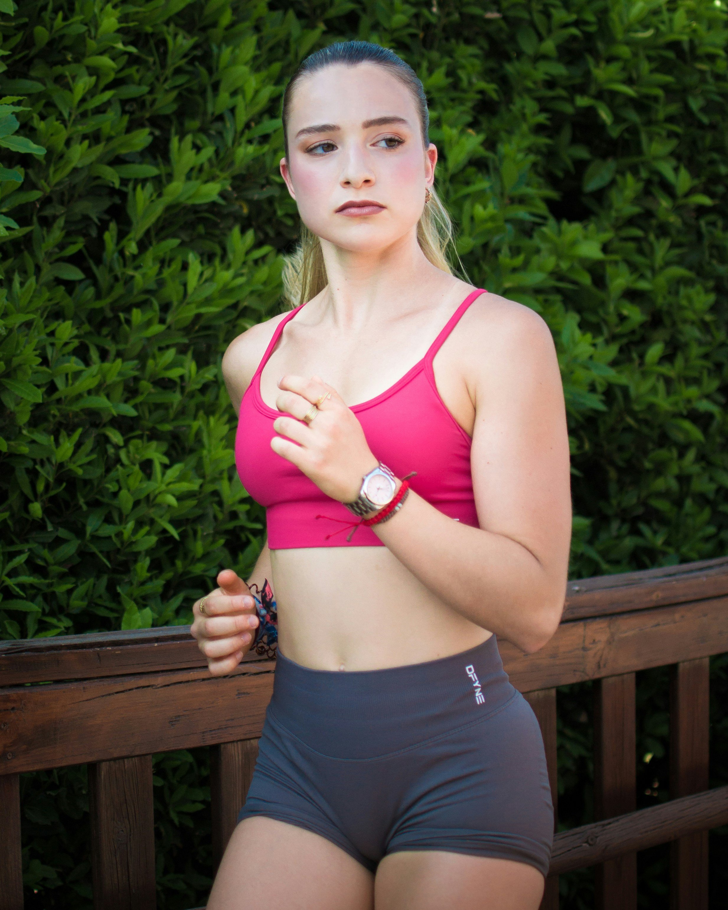 A young woman in athletic wear running outdoors, wearing a pink sports bra and dark shorts, with a background of green foliage and a wooden railing.