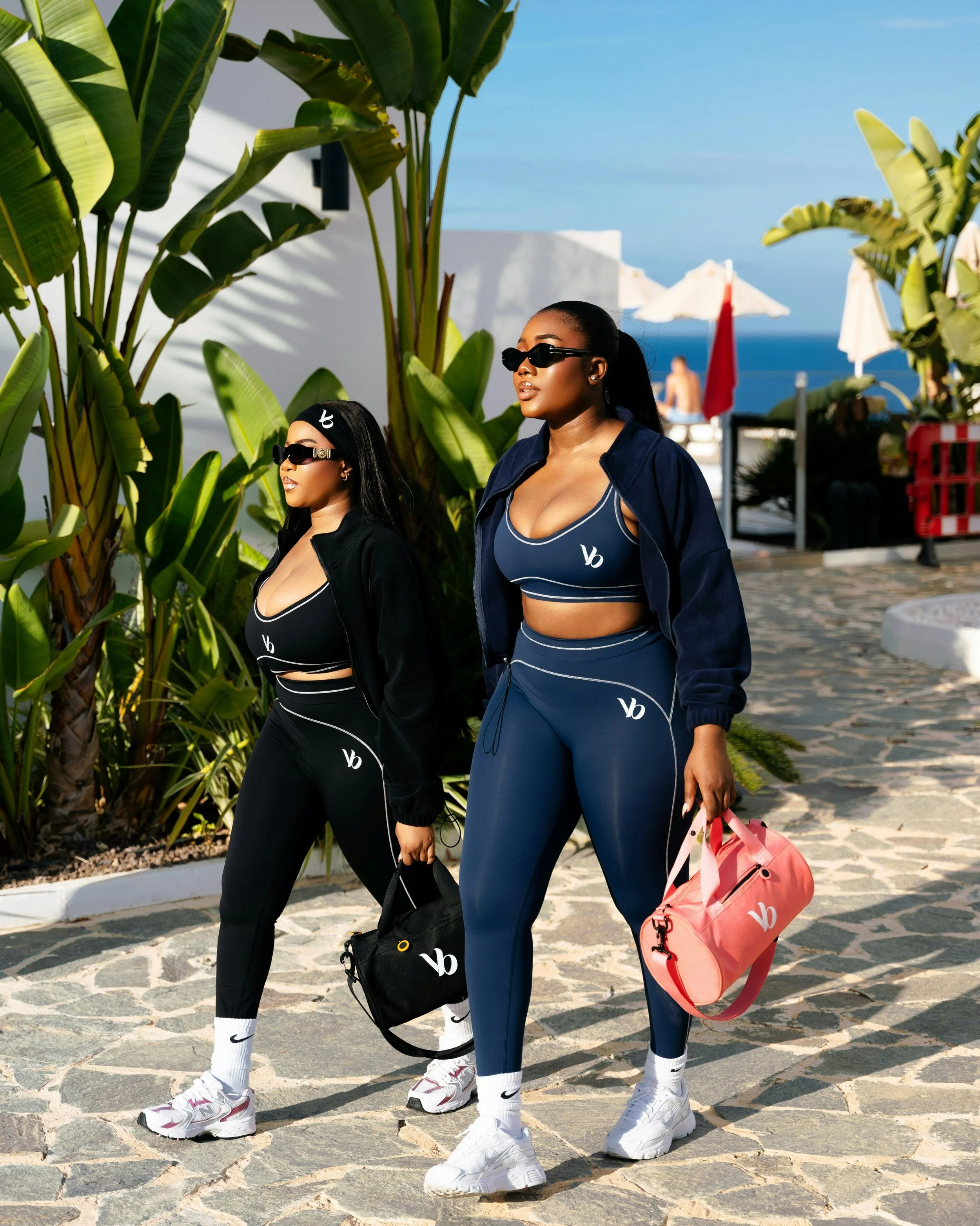 Two women walking outdoors near large green tropical plants, with white buildings and blue sky in the background. They are dressed in matching navy sportswear with white accents and logos, wearing sunglasses, and carrying sneakers and duffel bags.