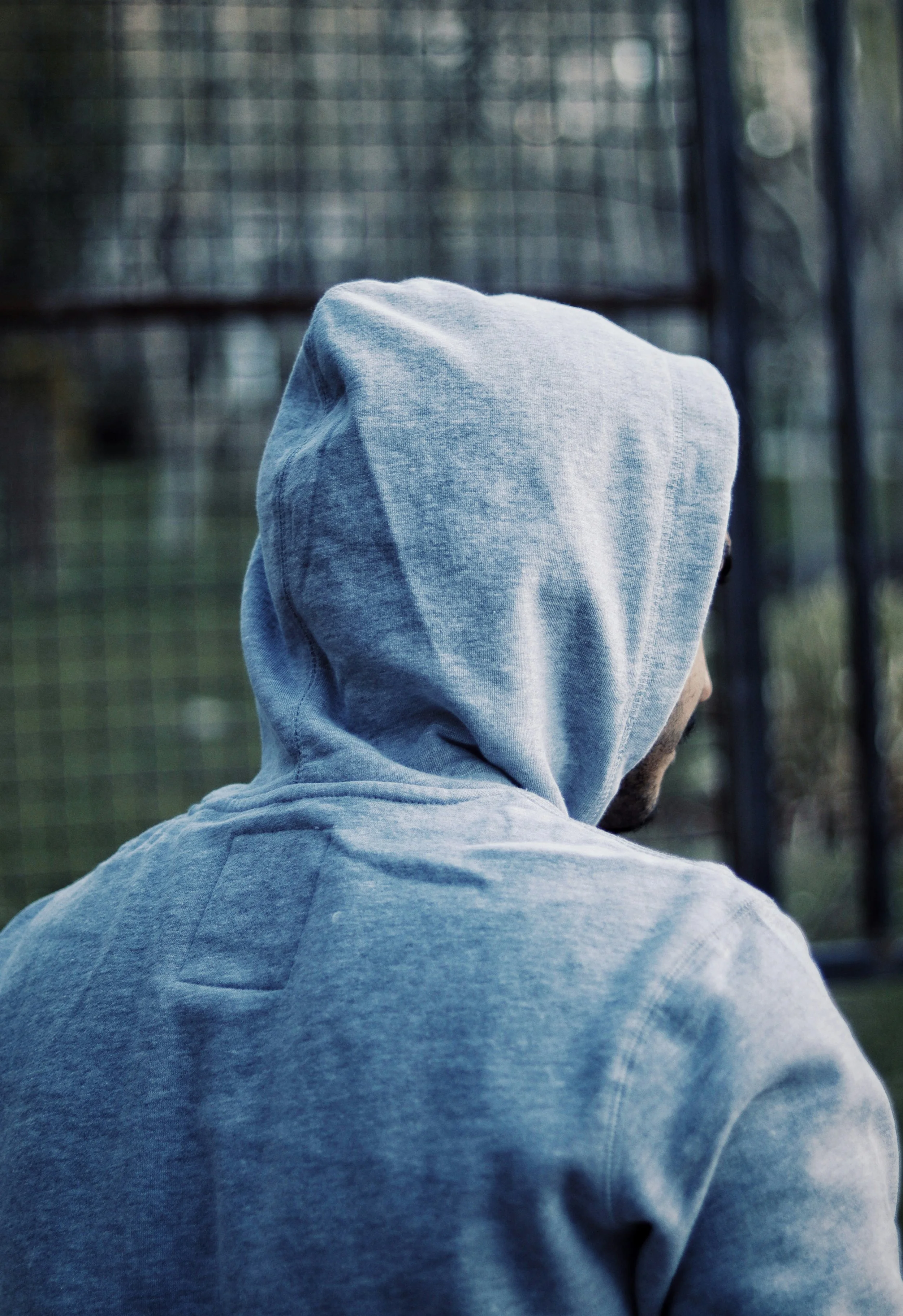 A person wearing a gray hoodie, viewed from behind, standing outdoors near a metal fence with blurred background.