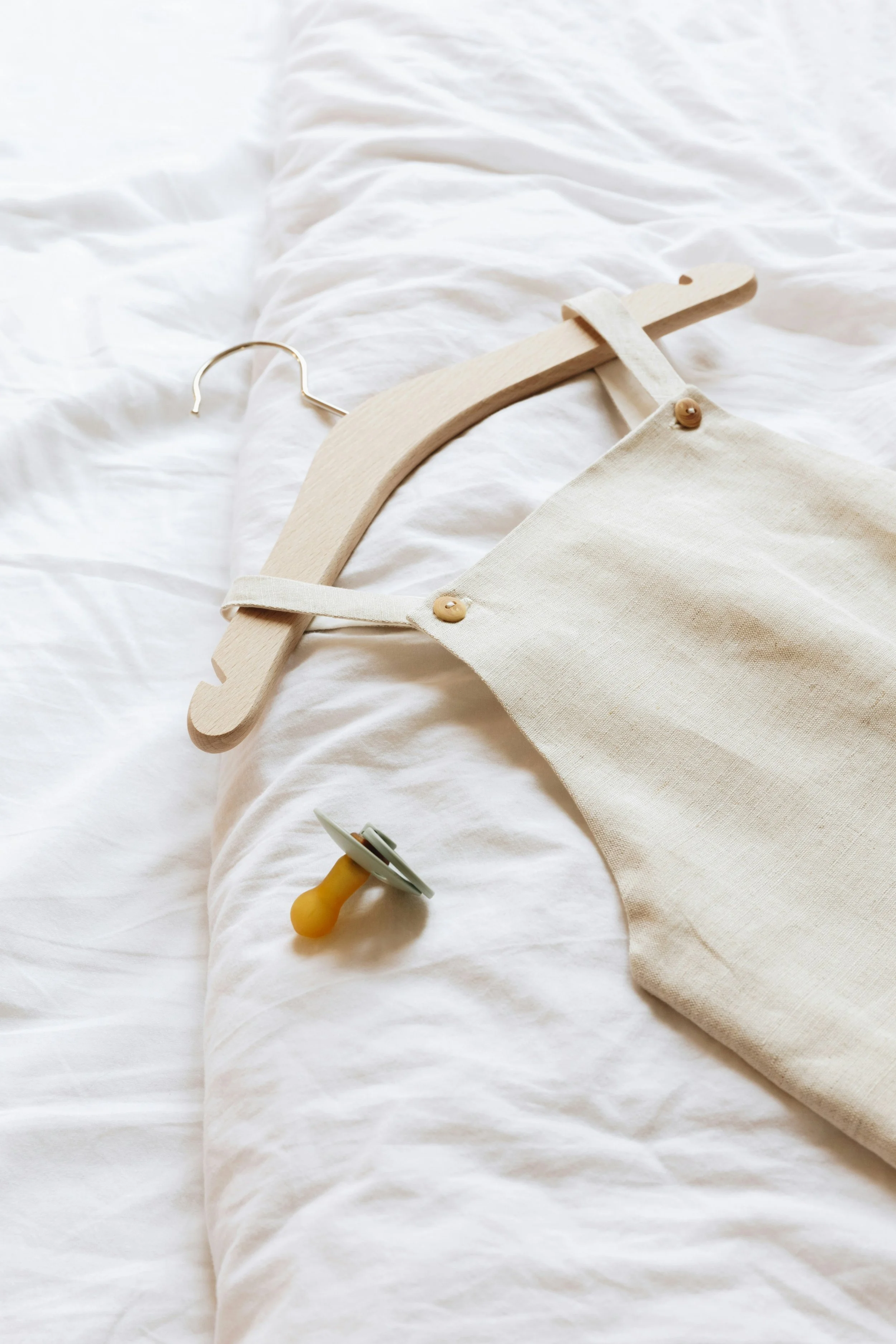 A white fabric tote bag on a white bed, hanging on a wooden hanger. A pacifier and a toy are also on the bed.