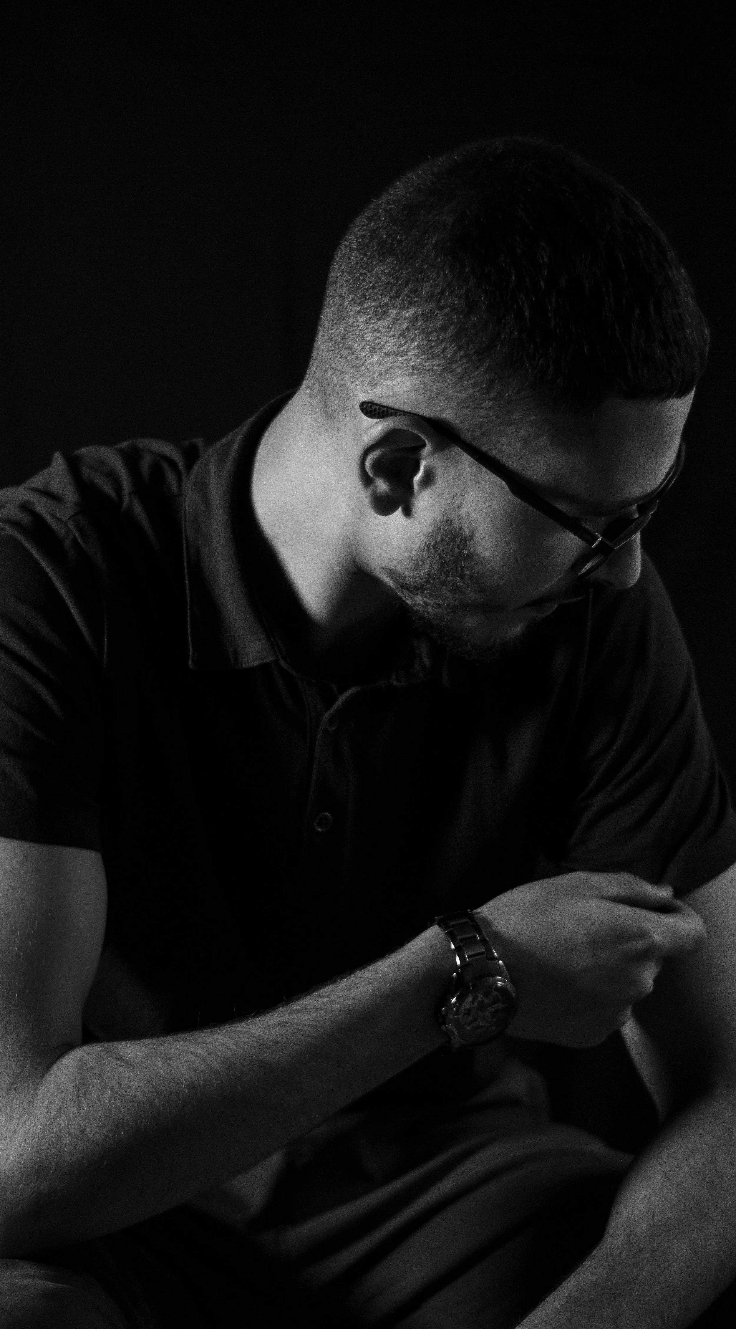 A young man with glasses and a wristwatch, wearing a dark polo shirt, sitting with his head slightly bowed and arms resting on his lap, captured in black and white against a dark background.