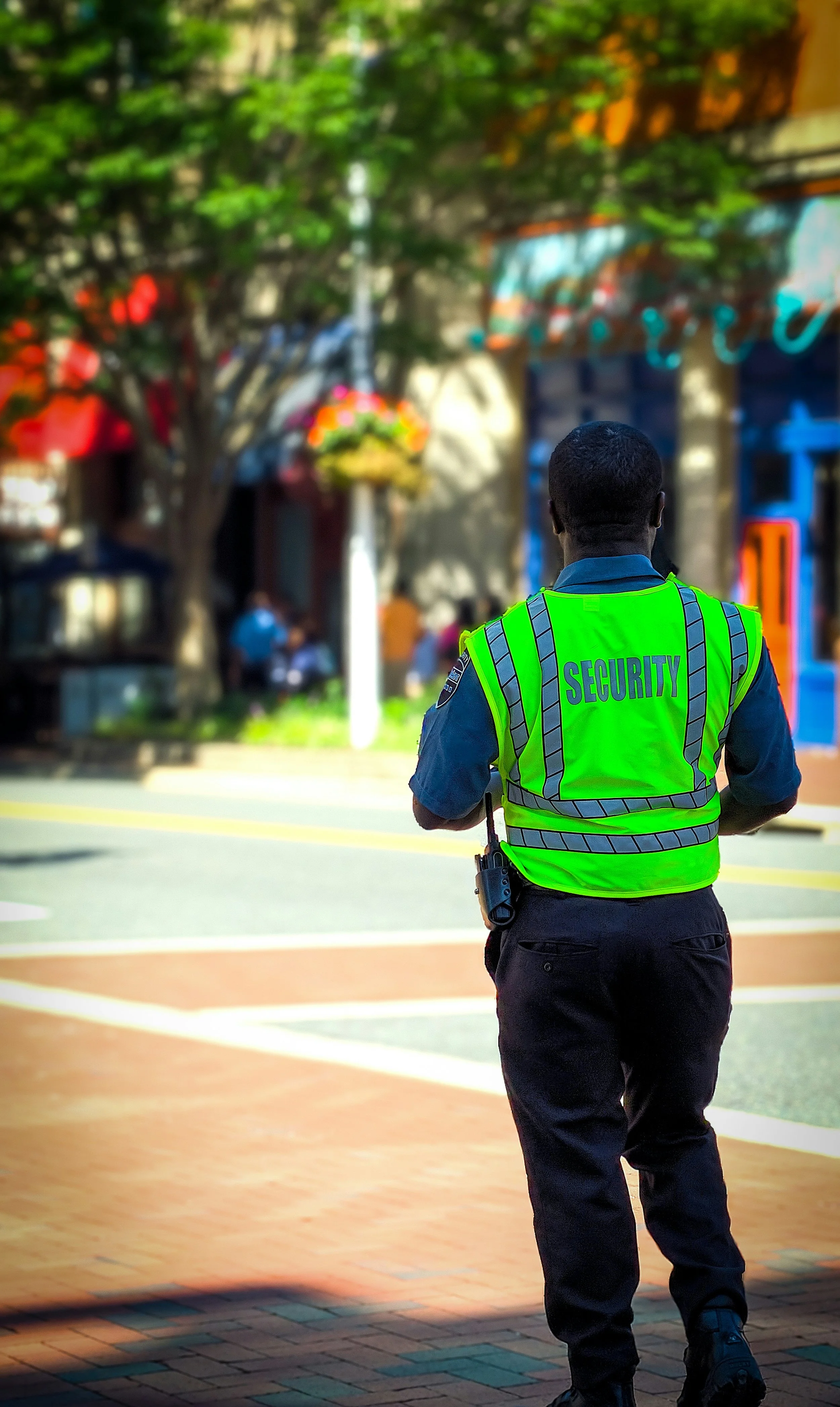 A security officer in uniform with a high-visibility vest that says 'SECURITY' on the back, standing outdoors on a brick-paved sidewalk, with trees and blurred buildings in the background.