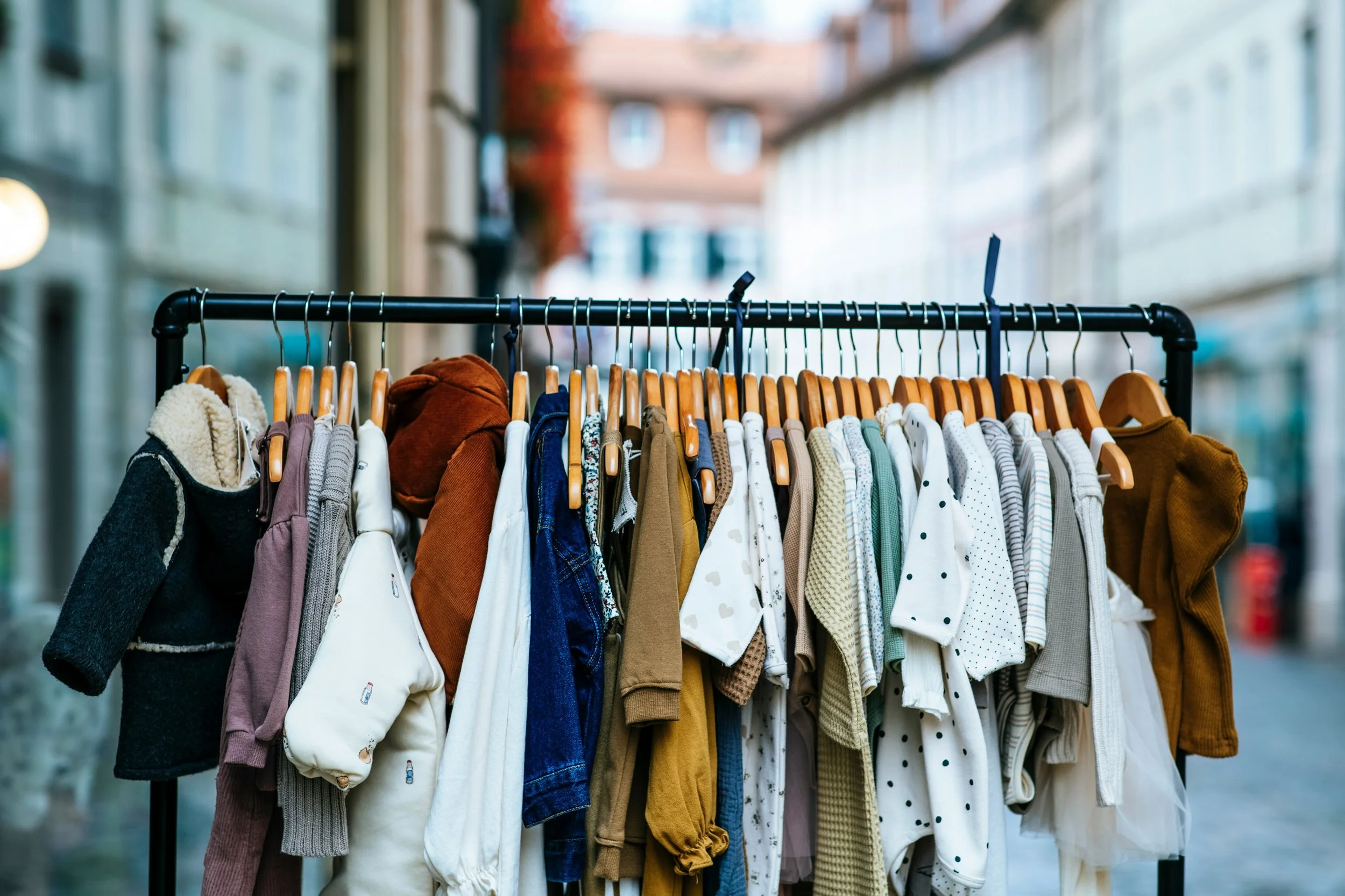 Clothing items hanging on a black clothing rack outdoors in an urban area with blurred buildings in the background.