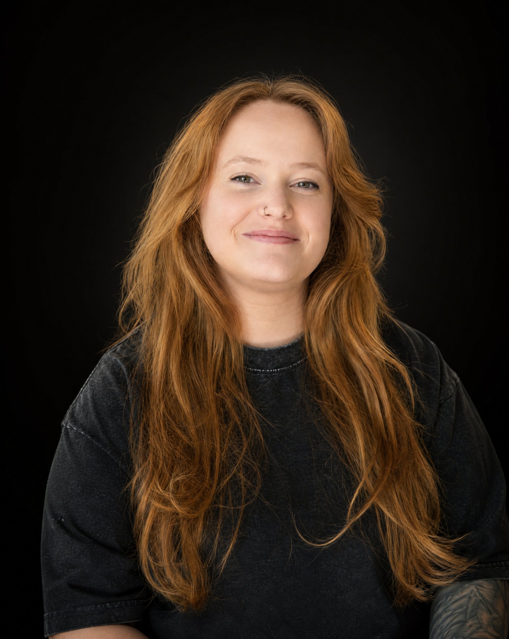 A young woman with long, wavy red hair, wearing a black and beige tie-dye shirt, smiling in front of a dark gray background.