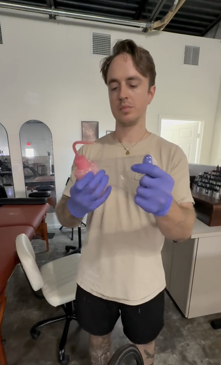 A tattoo artist, Mark Bennett "Dadtooer"  wearing a beige T-shirt and black shorts, holding a pink and a purple douching bulb in a room with tattoo salon chairs and shelves of tattoo ink.