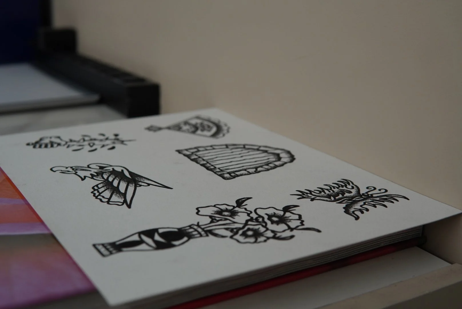 A sheet of paper with tattoo flash black line drawings of various objects, including a flower, a boat, a basket, a lantern, a plant, and a bird, resting on a surface near a wall.