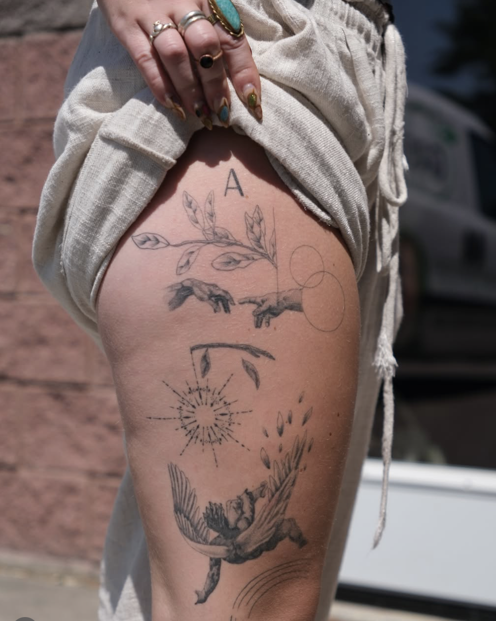 Black ink tattoo on person’s thigh featuring a bird with outstretched wings, a mythical creature, celestial and natural elements like leaves, a sunburst, and circles, with the letter A at the top.