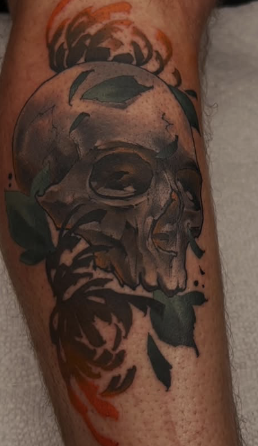 Tattoo of a human skull with leaf and flame accents on a person's leg.