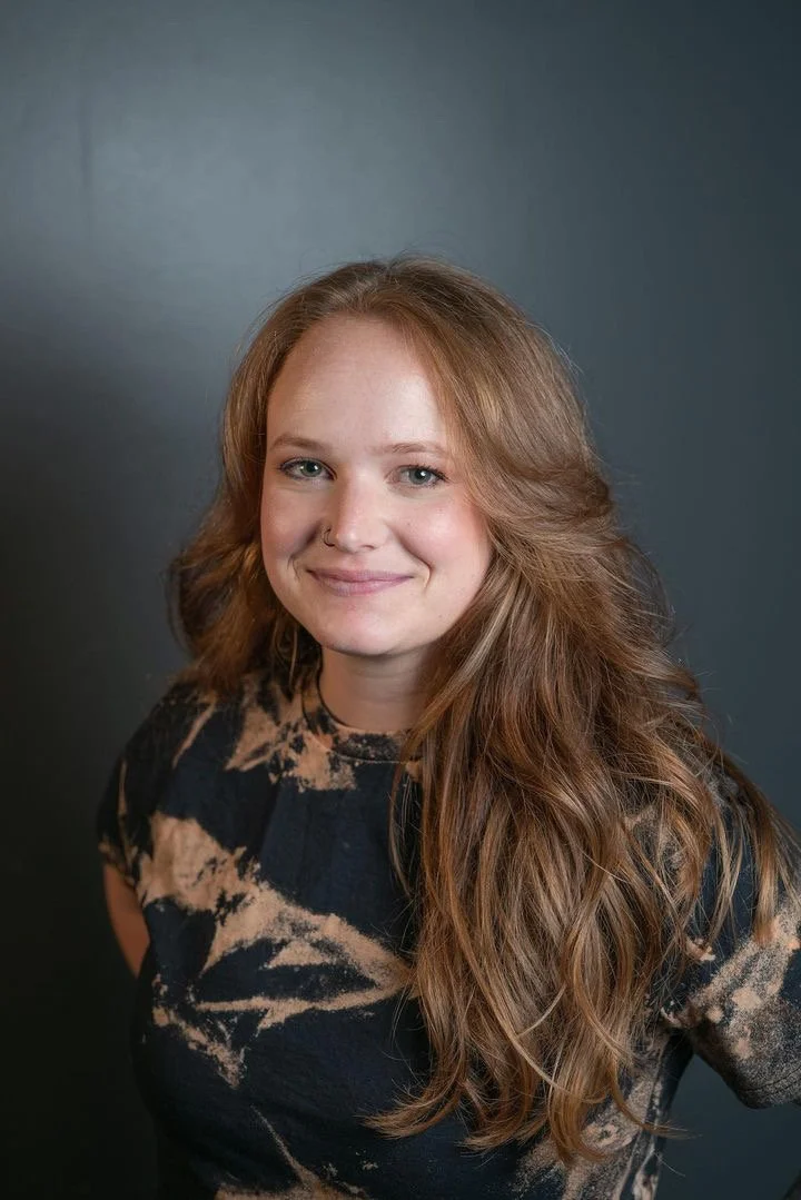 A young woman with long, wavy red hair, wearing a black and beige tie-dye shirt, smiling in front of a dark gray background.