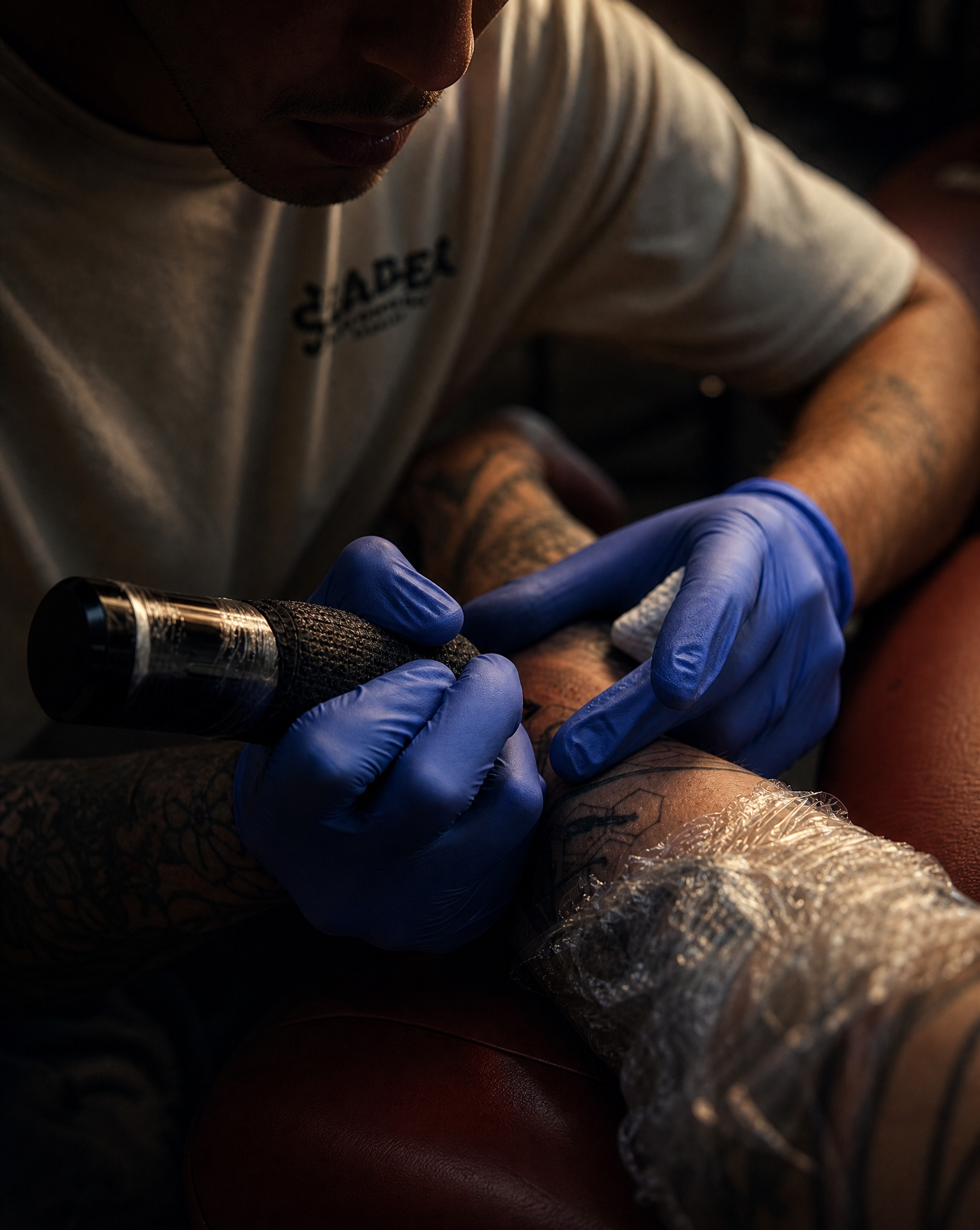 A tattoo artist wearing blue gloves is tattooing a person's arm, with focus on the artist's hand and the tattoo machine. The person receiving the tattoo has tattoos visible on their arm and is covered with plastic wrap.