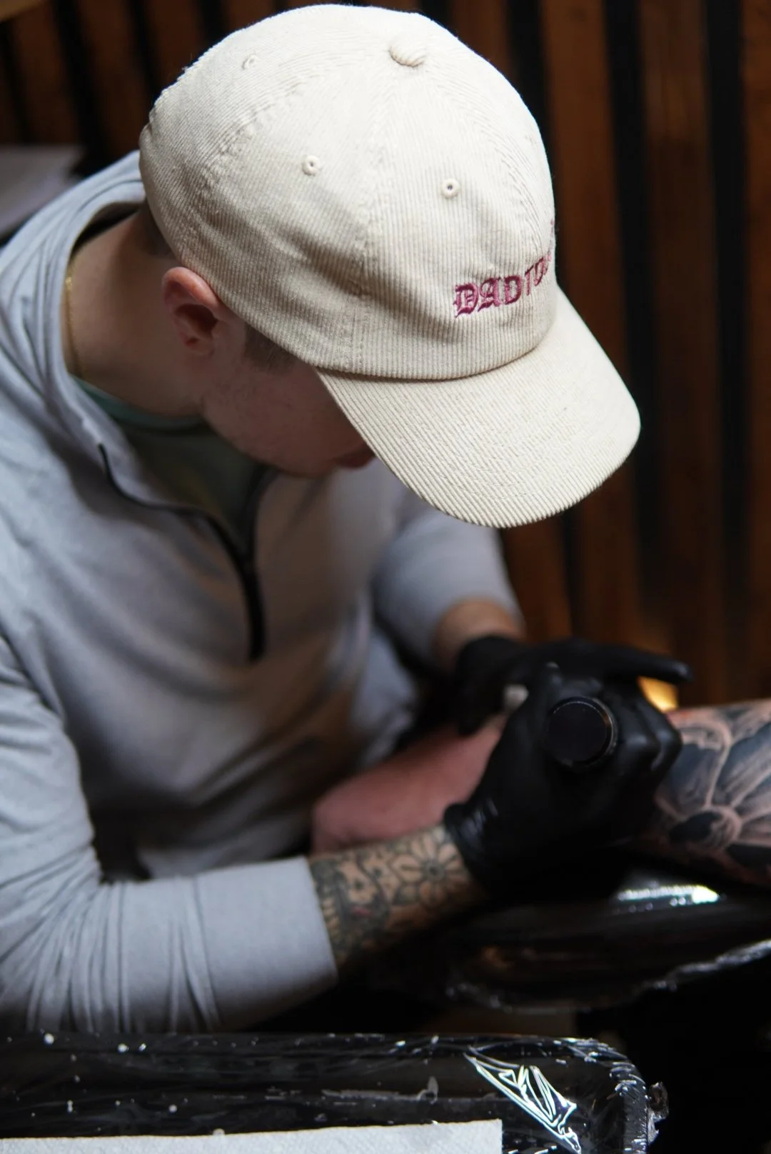 A person wearing a white baseball cap with the word 'DADDY' embroidered in red, a gray zip-up hoodie, and black gloves, working on a tattoo on their forearm.