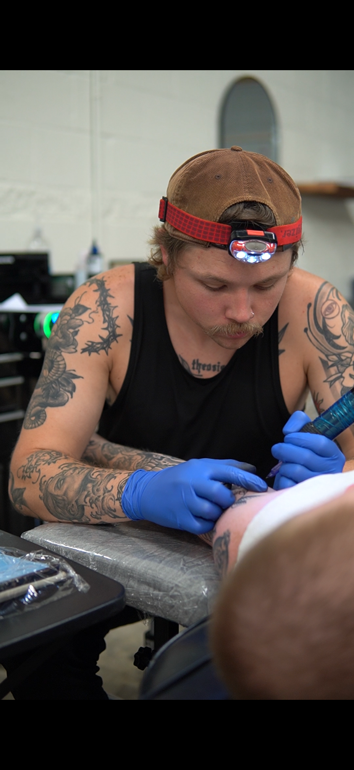 A tattoo artist with tattoos on his arms and chest, wearing a brown cap with a headlamp, tattooing a person on their leg in a tattoo studio.