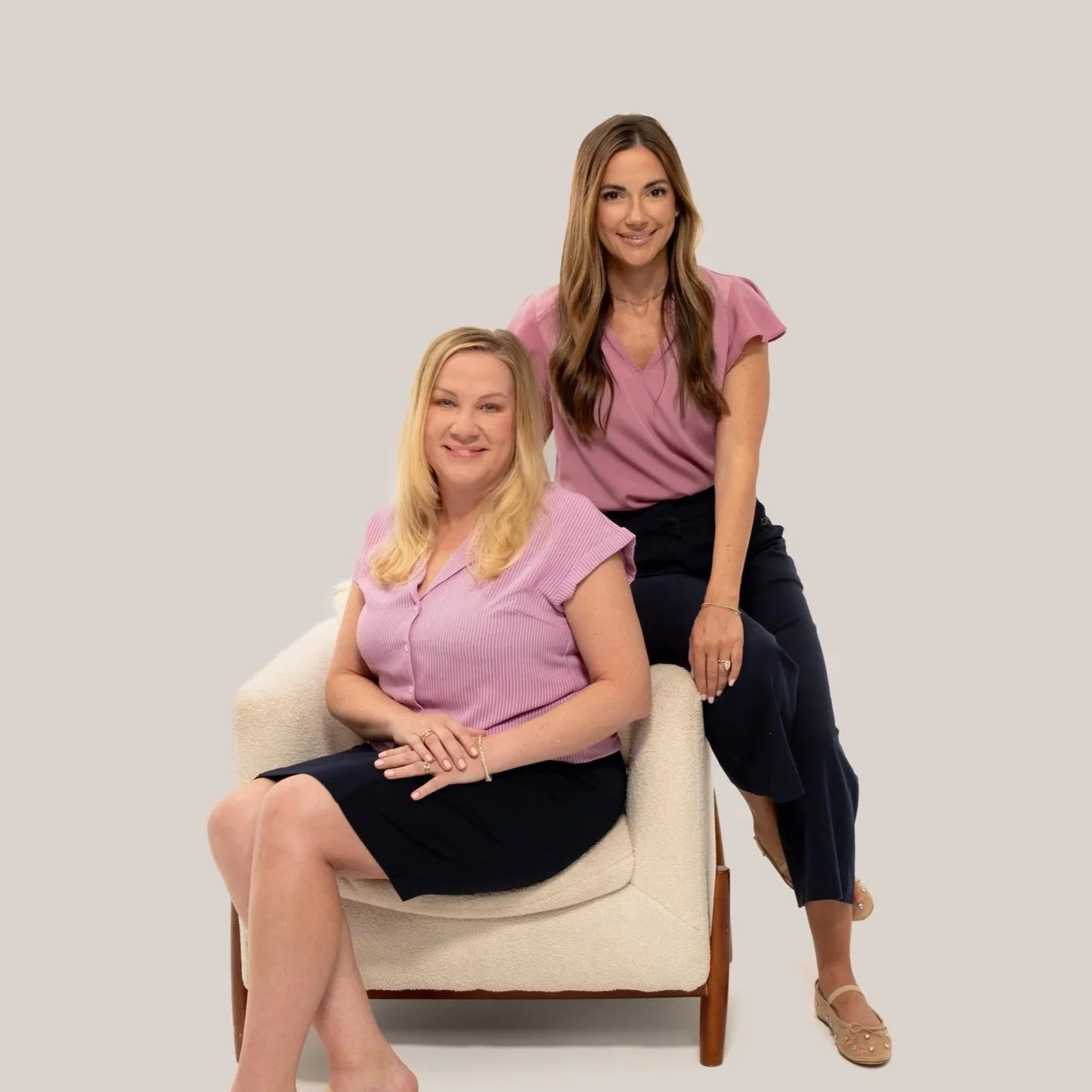 Founders of Arcadia Women's Wellness, Julia Cyr, DNP and Kristina Calligan, FNP posing together against a neutral background. Kristina is seated in a chair, smiling, and Julia is leaning.
