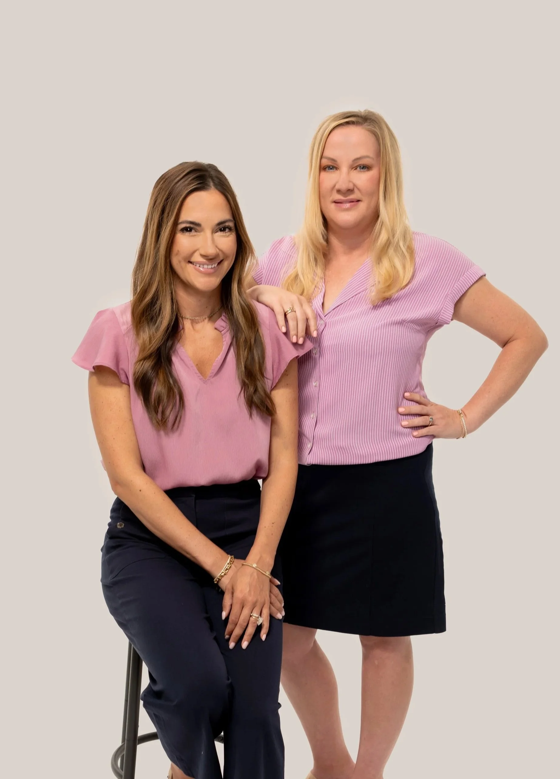 Founders of Arcadia Women's Wellness, Julia Cyr, DNP and Kristina Calligan, FNP posing together against a neutral background. Julia is seated on a stool, smiling, with long wavy brown hair, and Kristina is standing.