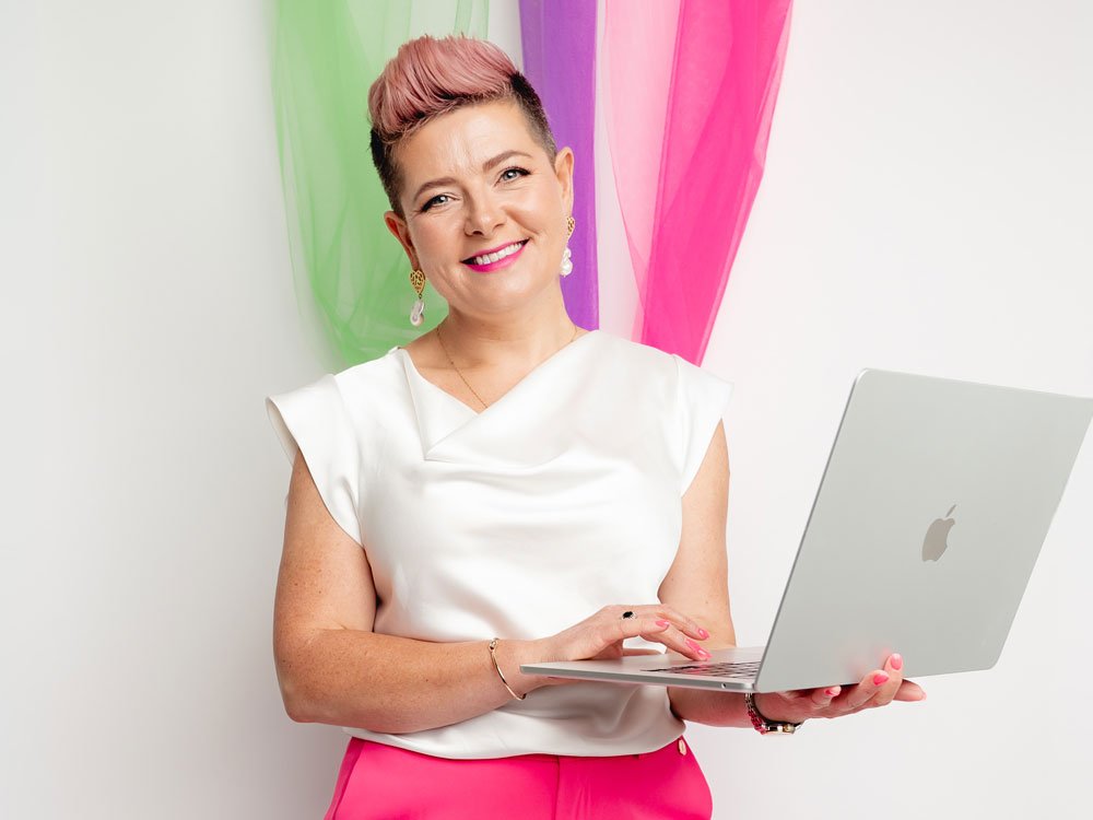 Ciara Crossan smiling while holding a silver laptop, wearing a white sleeveless top, bright pink trousers, pearl drop earrings, and pink quiff hairstyle with sheer green, purple, and pink drapes behind her.