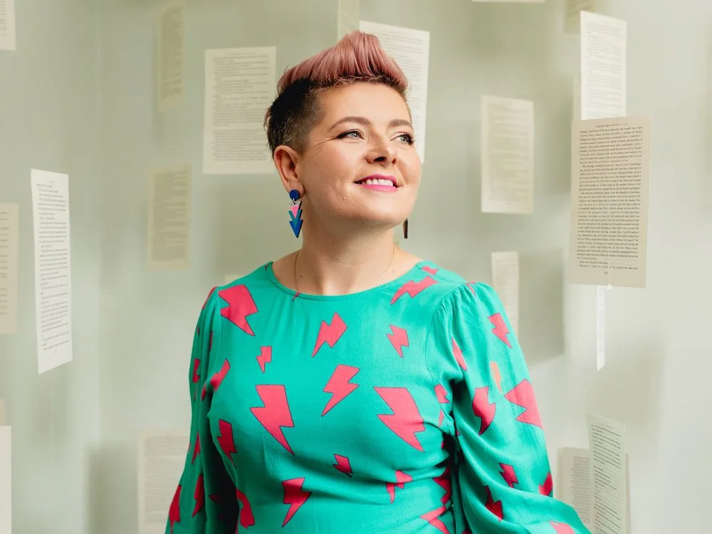 Ciara Crossan smiling and looking upward in a teal dress with pink lightning bolts, pink quiff hairstyle, and blue arrow earrings, surrounded by suspended text pages.