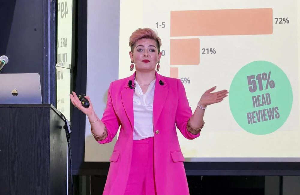 Ciara Crossan giving a keynote in a bright pink suit, gesturing in front of a presentation slide with survey results, including “51% read reviews.”