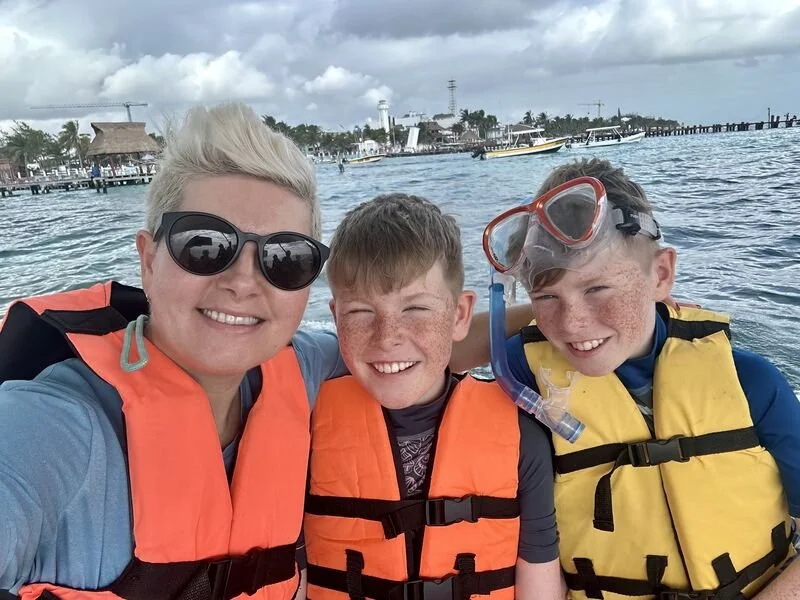 Ciara and her boys wearing orange and yellow life jackets on a boat, with one boy in snorkel gear and a marina behind them under cloudy skies.