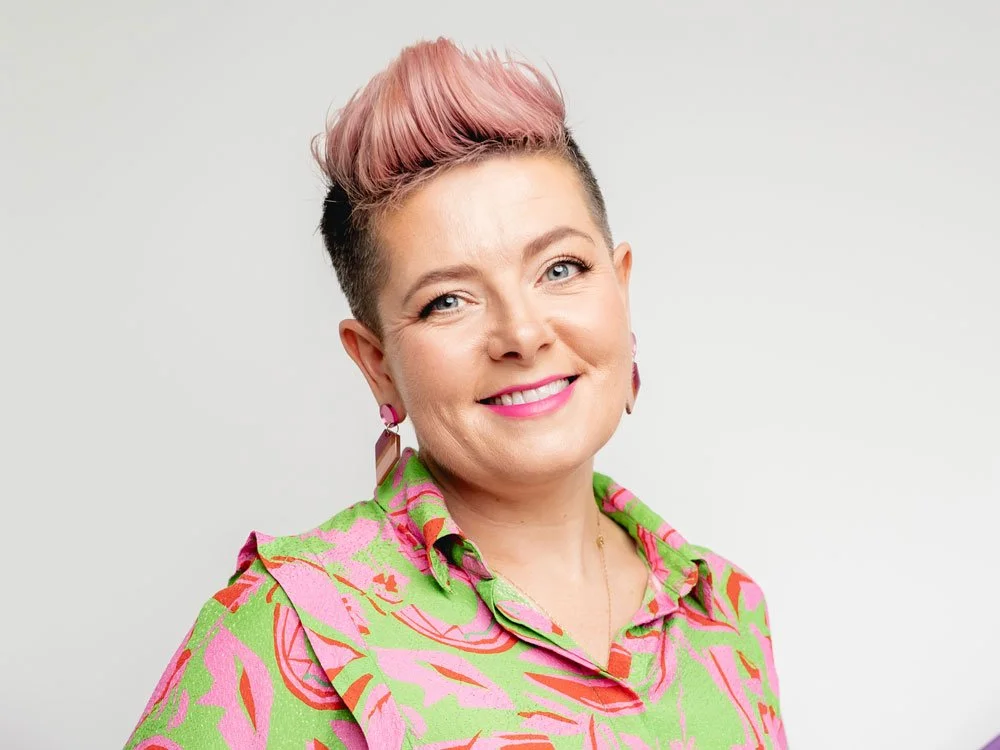 Ciara Crossan smiling in a green and pink patterned shirt, with pink quiff hair, bright earrings, and pink lipstick against a plain light background.
