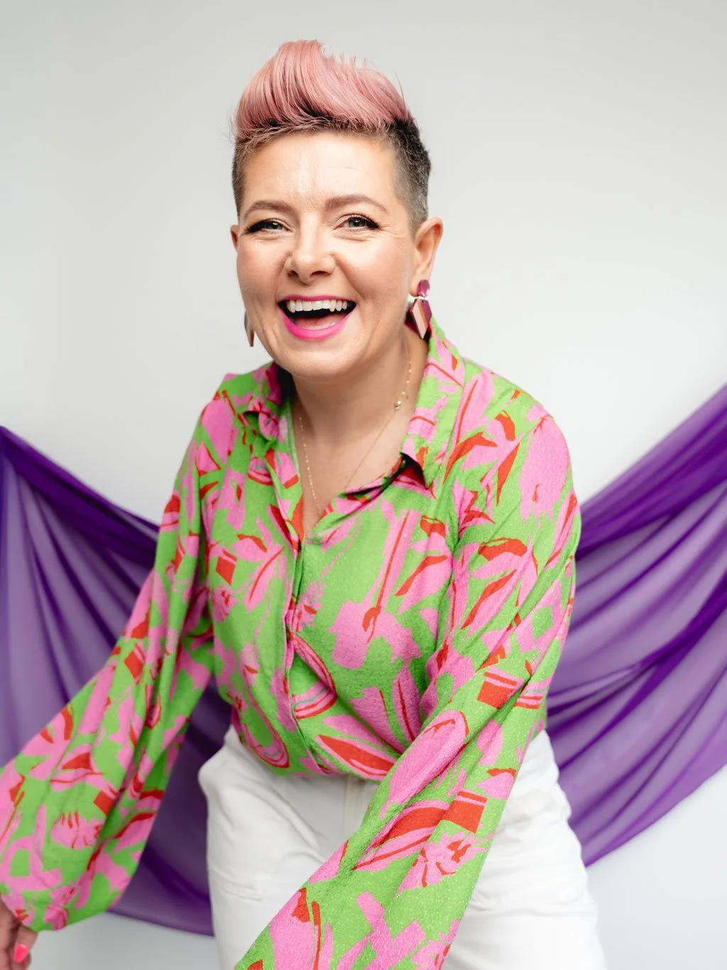 Ciara Crossan laughing in a green and pink patterned shirt with white trousers, pink quiff hairstyle, and bold earrings, leaning forward in front of purple draped fabric.