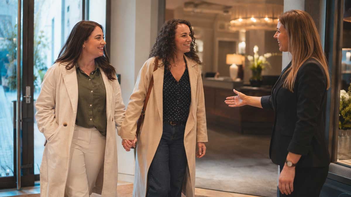 Two women holding hands are warmly greeted by a staff member in a bright hotel lobby or reception area.