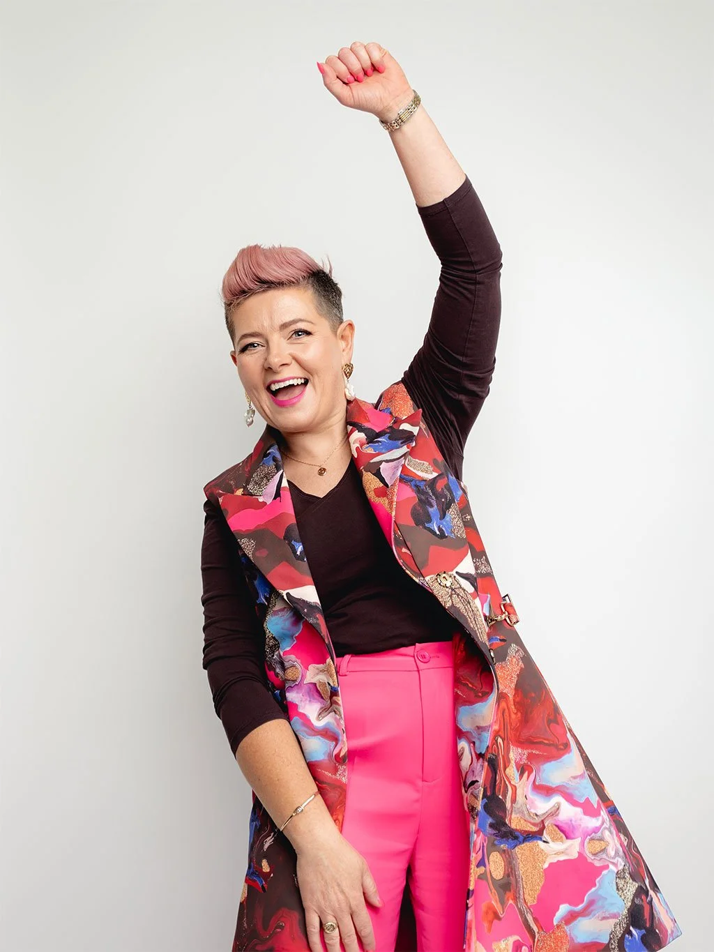 Ciara Crossan smiling with one arm raised, wearing hot pink trousers, a dark top, and a colourful patterned sleeveless coat against a plain light background.