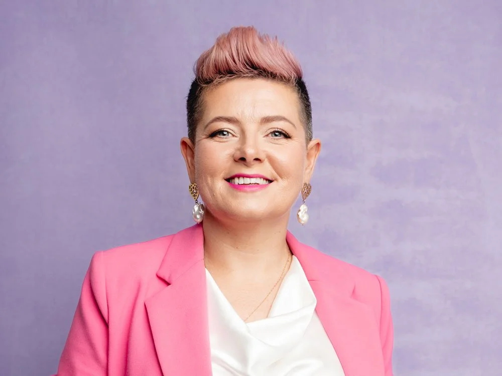 Ciara Crossan smiling in a pink blazer over a white top, with pink quiff hair and pearl drop earrings, against a soft purple background.